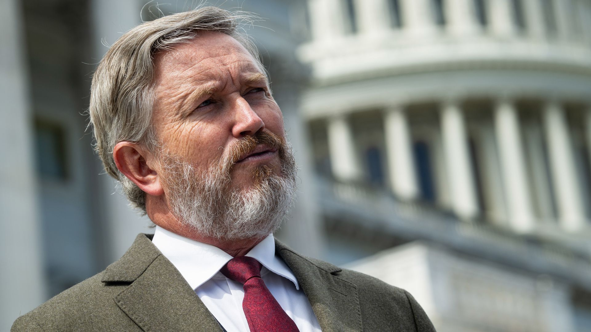 Close-up of an older man with gray hair and beard, wearing a brown suit and red tie, gazing upward. A pale, columned building is blurred in the background.