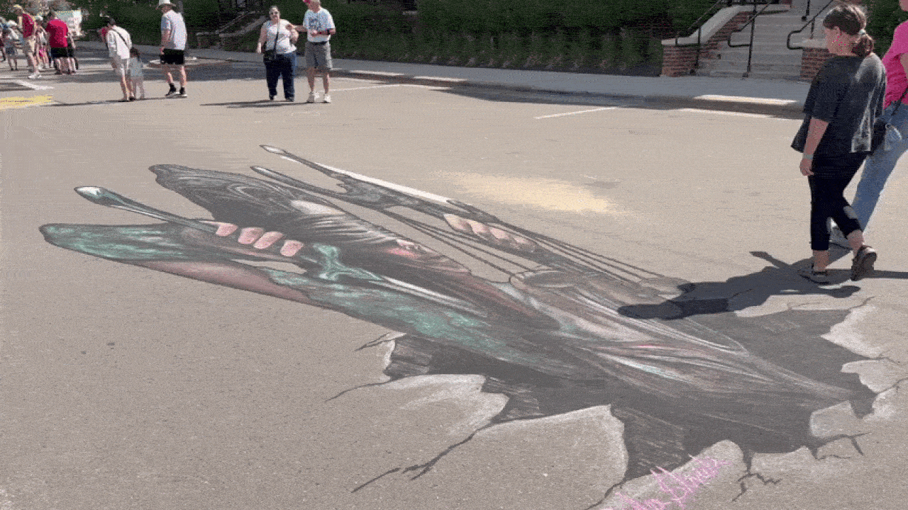 Large 3D chalk butterfly drawn on a parking lot, with dark wings and pink-green accents; several people walk nearby, casting shadows on the pavement.