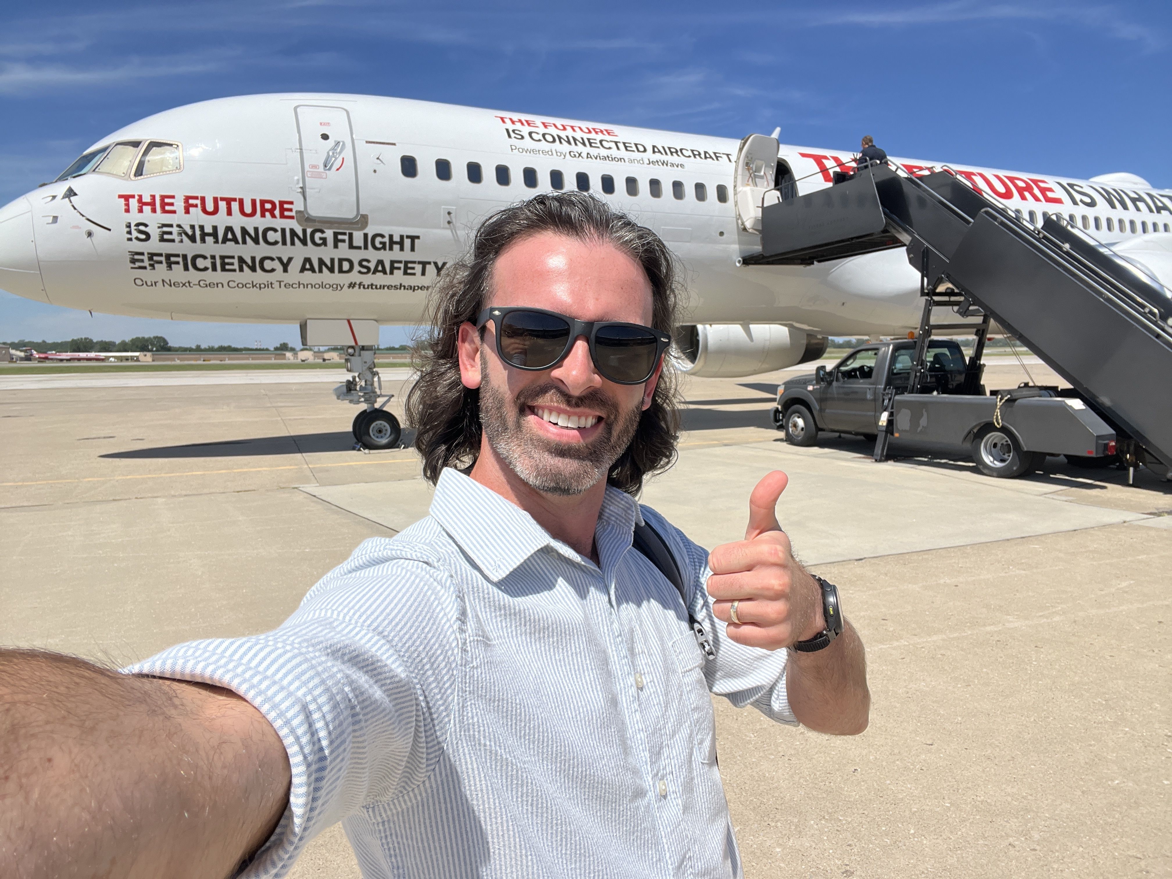 Man with sunglasses and long hair takes a selfie, giving a thumbs-up in front of a white airplane with red and black text about future flight efficiency and connected aircraft, on a sunny tarmac.