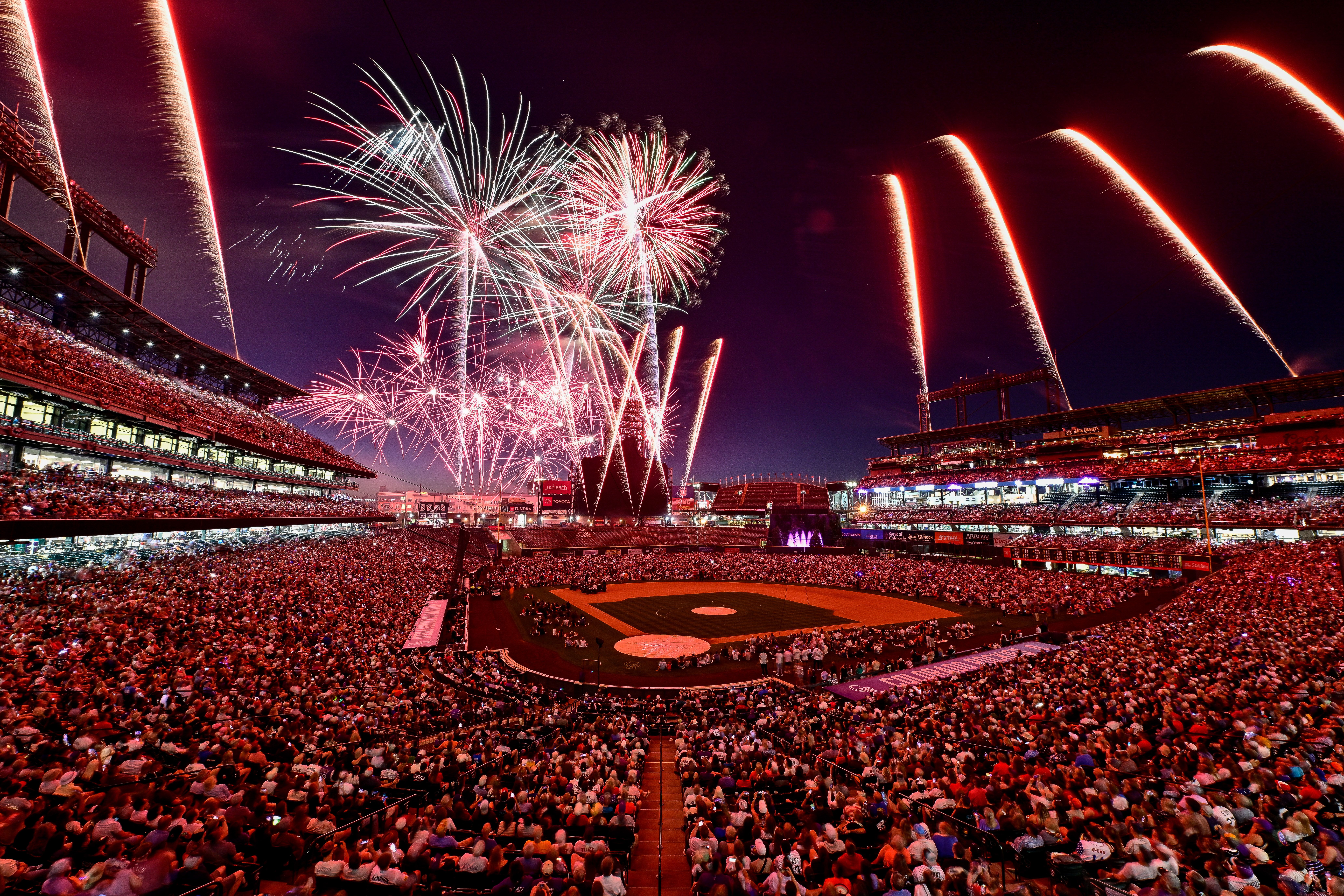 Nighttime stadium filled with a cheering crowd as red and white fireworks fly over the baseball field, lighting the grandstands on both sides.
