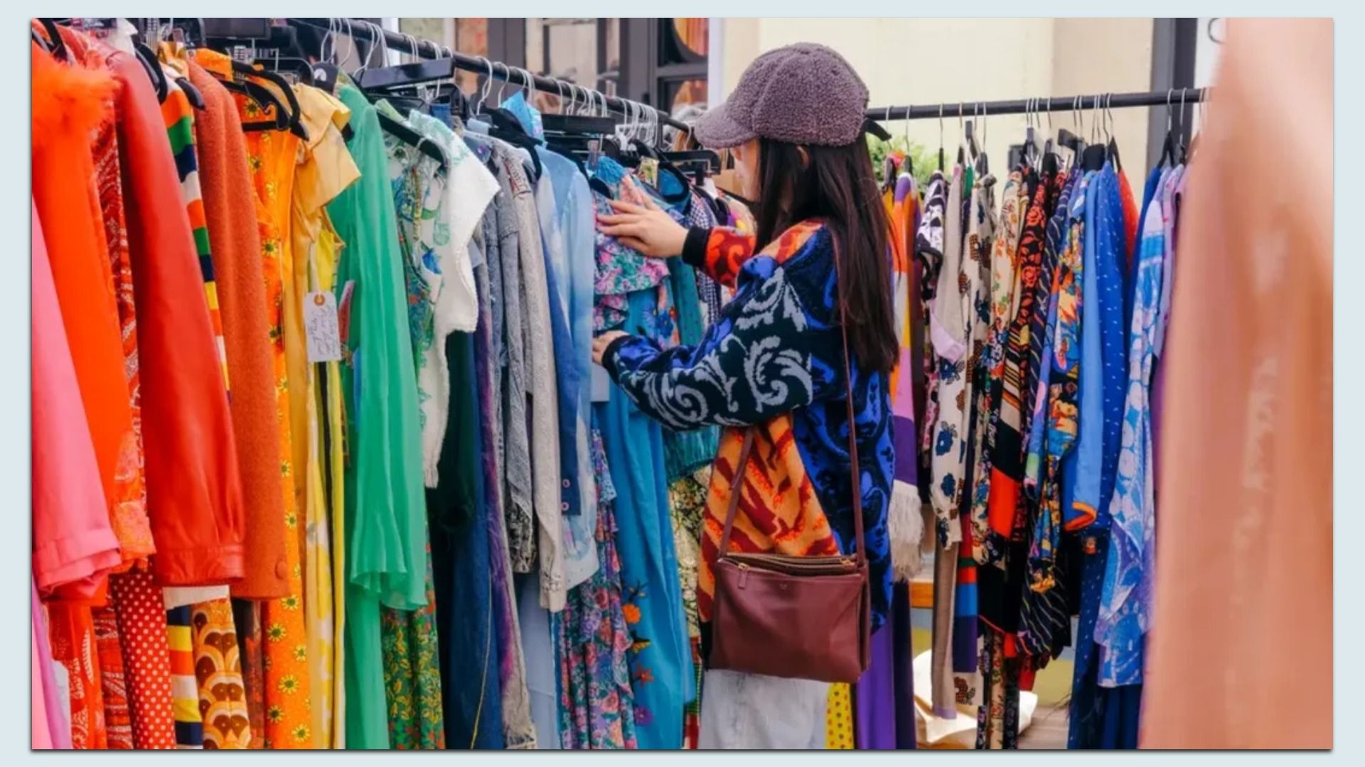 A person wearing a brown fuzzy cap and a patterned blue jacket with a brown crossbody bag browses a rack of colorful dresses in reds, oranges, greens, blues at a clothing shop.