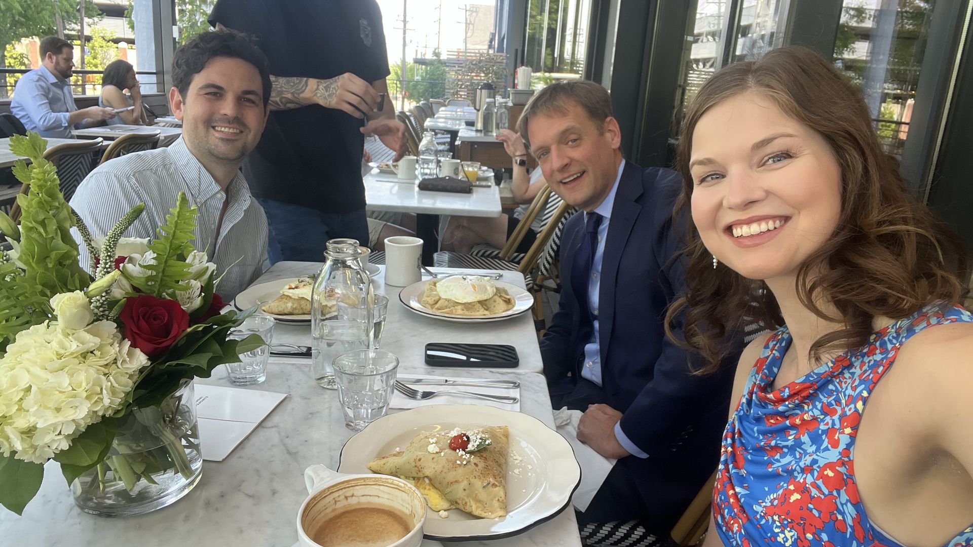 Selfie of three friends at an outdoor cafe: woman in a blue floral dress smiles at the camera; two men sit at a marble table (one in a suit), with a waiter behind. Flower centerpiece and breakfast items on the table.