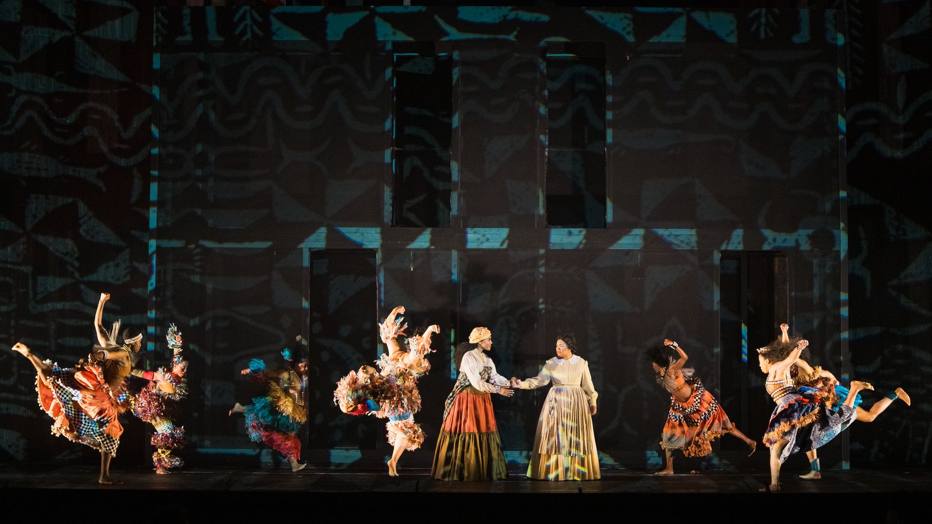 Theatrical stage scene with performers in colorful, patterned costumes and two central characters in period dresses holding hands against a dark background with tribal-inspired blue patterns.