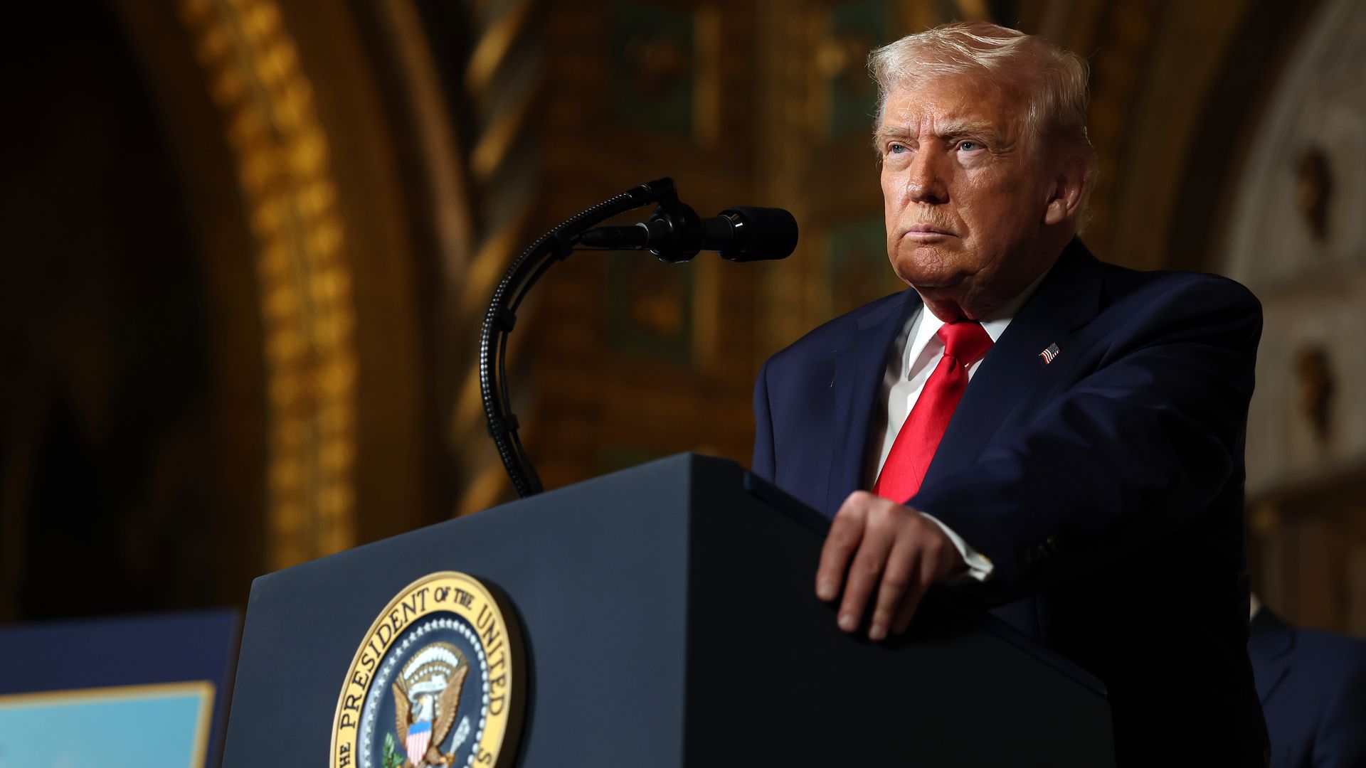 President Trump, wearing a navy jacket with a US flag pin at the top of his left lapel, white shirt and red tie, stares off as he stands at a blue podium saying "President of the United States."