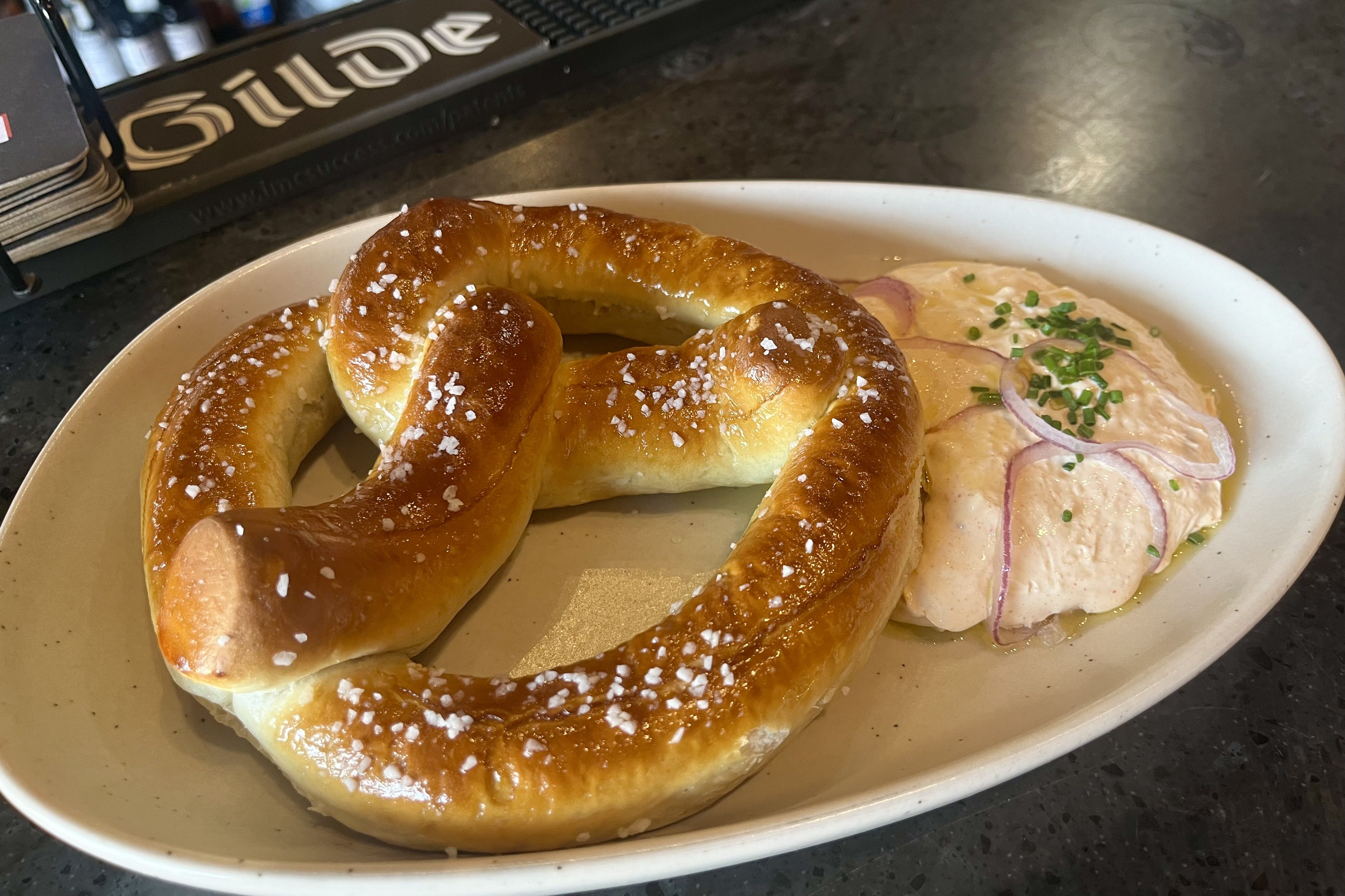 Golden-brown pretzel with coarse salt rests on a plate beside pale pink sliced meat, red onion rings, and chopped chives in a light sauce.