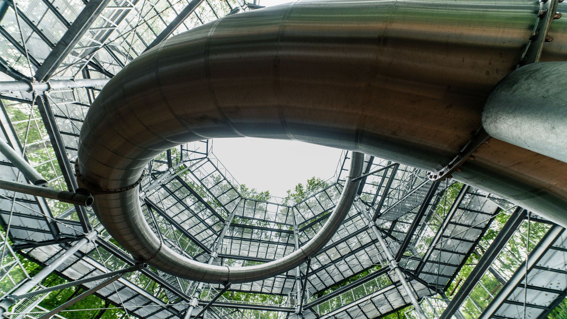 Upward view of a large, curved metal slide enclosed by a multi-level steel and glass structure with trees visible through the transparent panels.