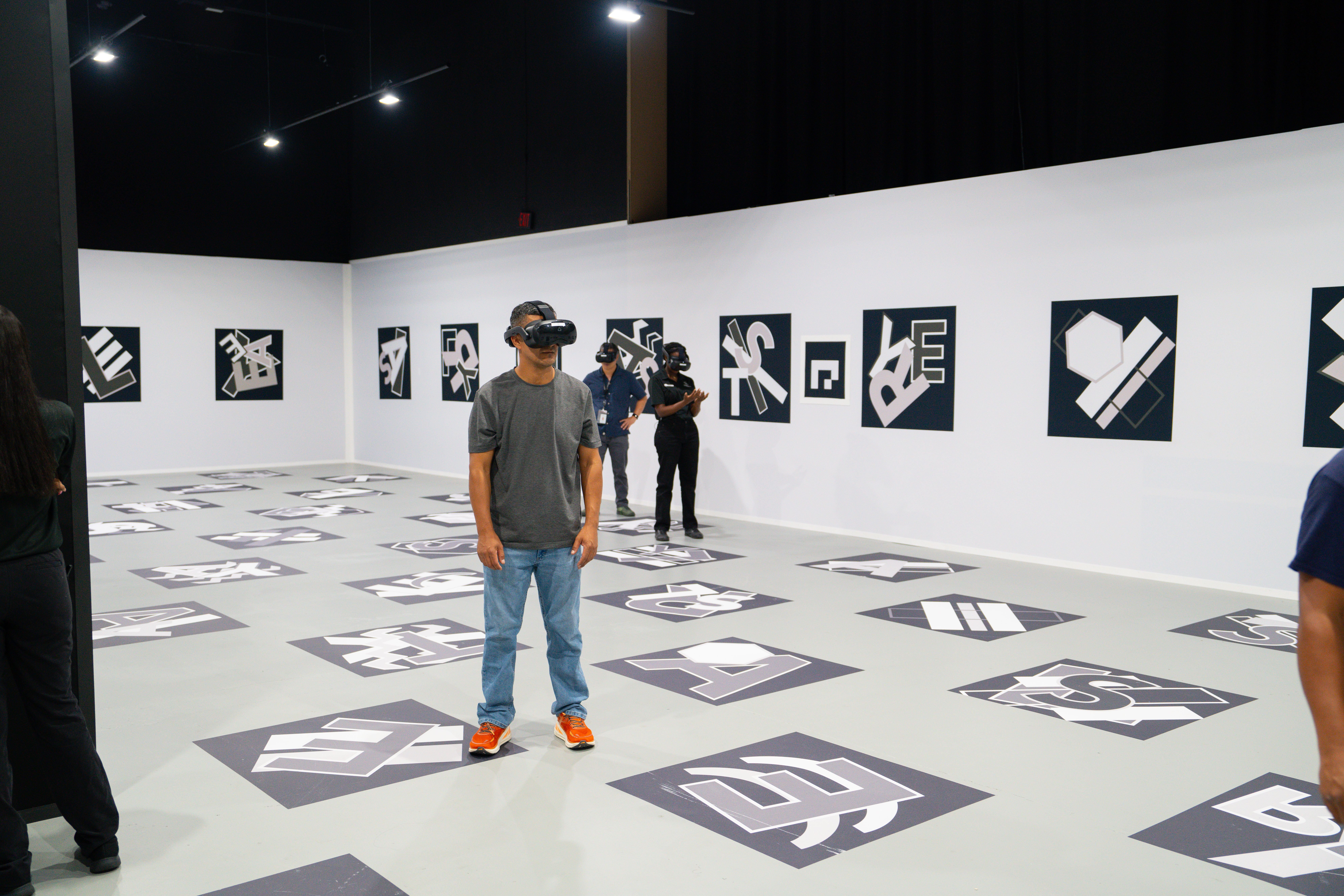 Man in a gray shirt and orange sneakers wearing a VR headset stands in a white-walled gallery. Black upper walls, geometric logos on wall panels and floor tiles around him, others in background.