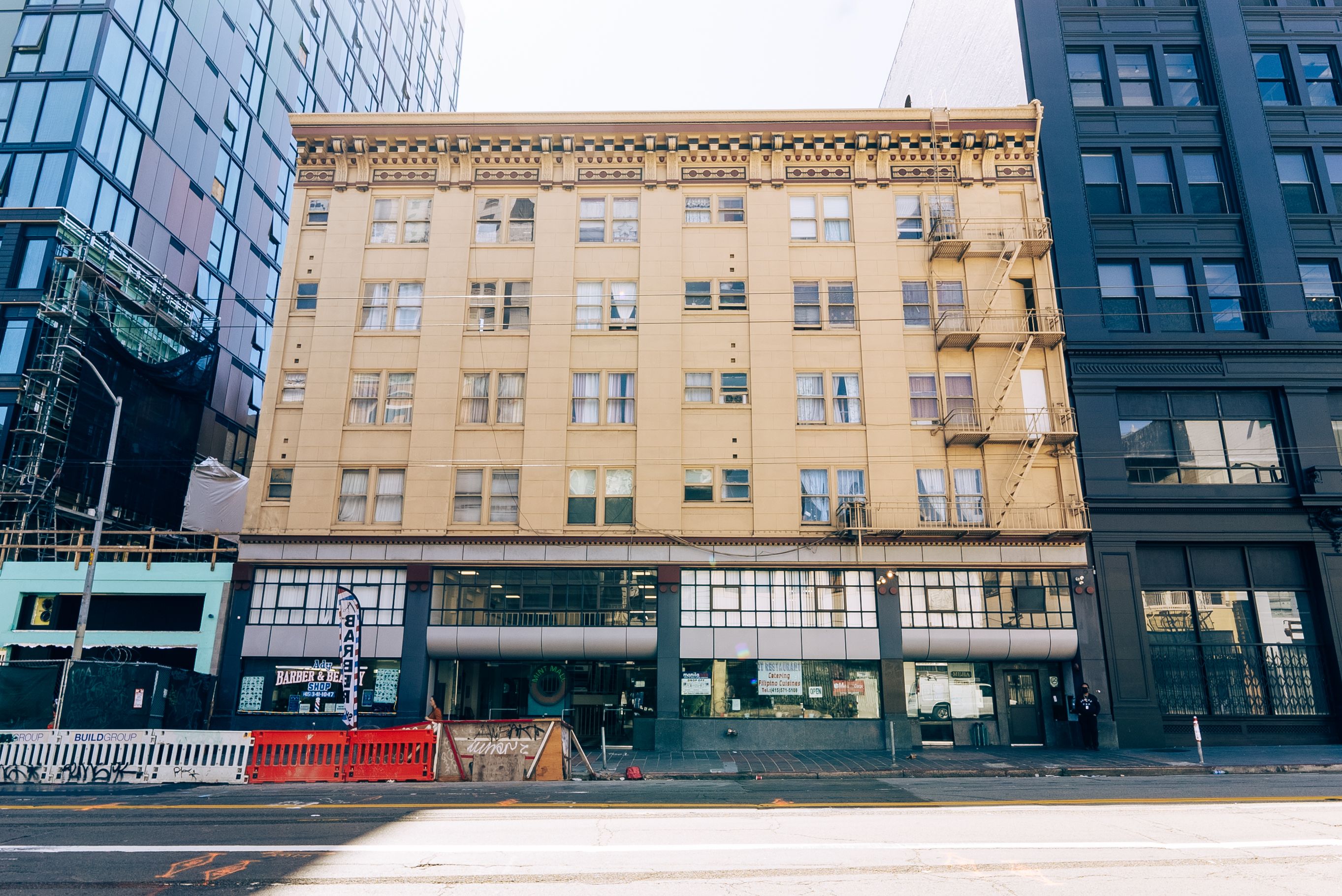 Street view of a beige five-story building with multiple windows and a fire escape, flanked by taller modern buildings in blue and black glass.