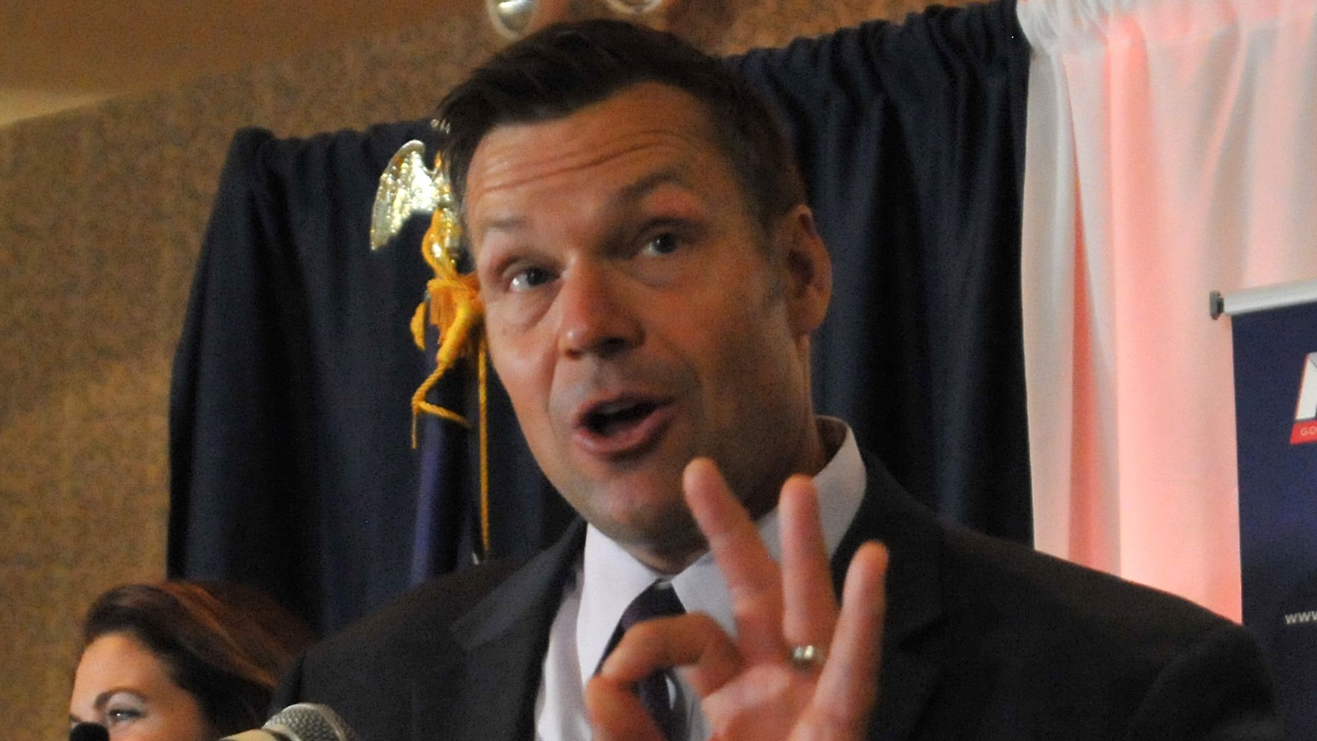 Kris Kobach speaking at a microphone holding up the "okay" sign with his left hand