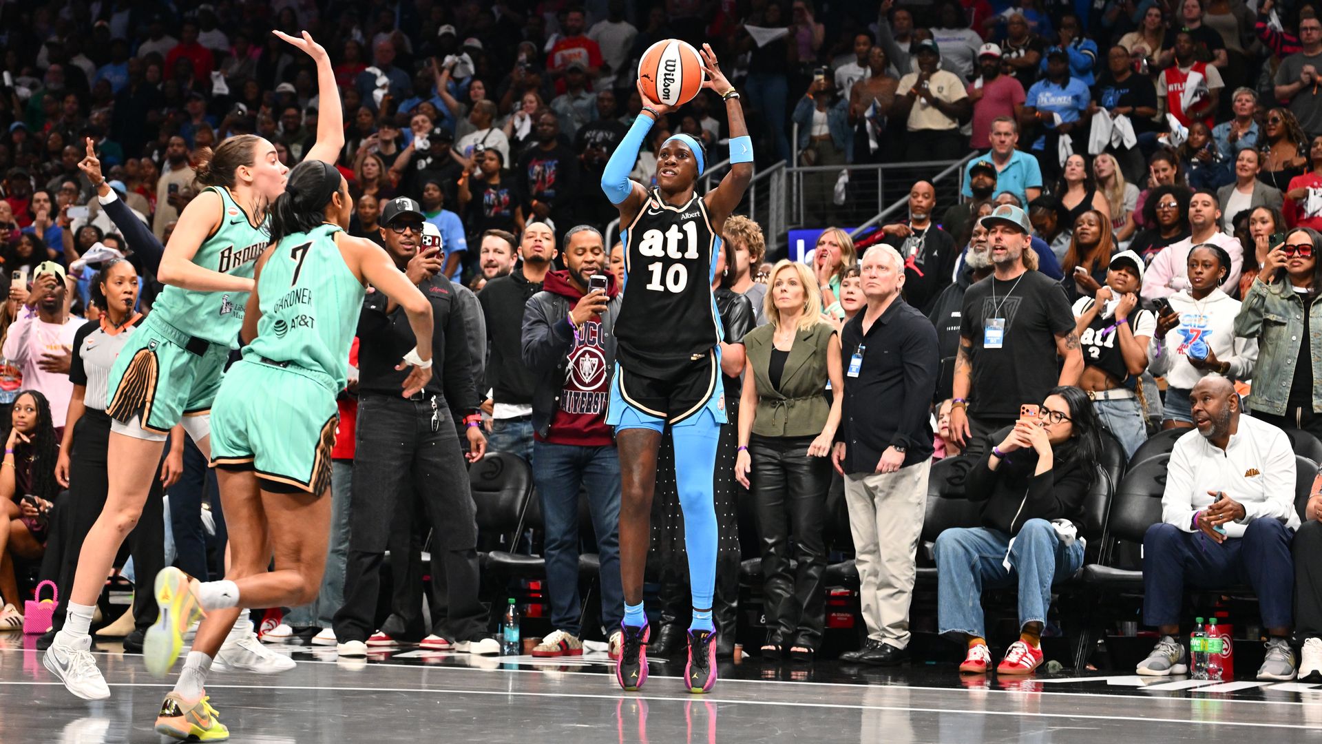 A player wearing a black uniform with the letters "ATL" and number "10" on the front and blue compression sleeves on the left leg and right arm holds a basketball in the air on a court. Two players in light green uniforms are seen on the court and fans watch in the background.