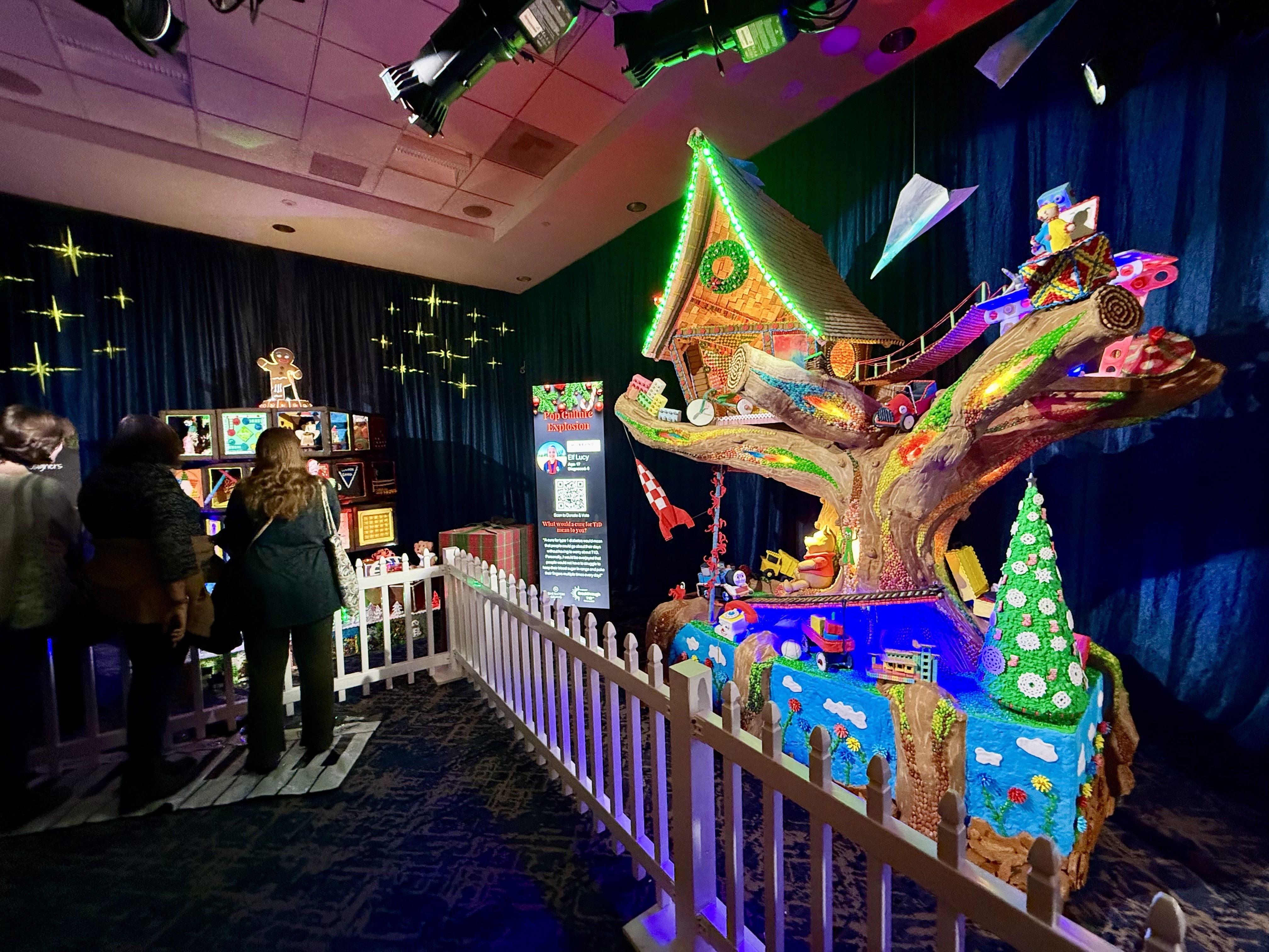 Colorful, detailed gingerbread treehouse display with a lighted treehouse, toys, and a decorated Christmas tree, surrounded by a white picket fence, with people observing in dim lighting.