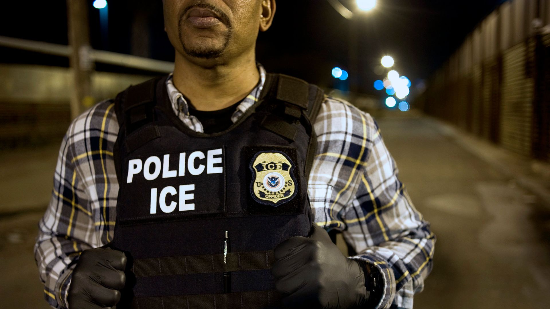 In this image, an ICE officer holds onto his vest as he stands in the street at night. 