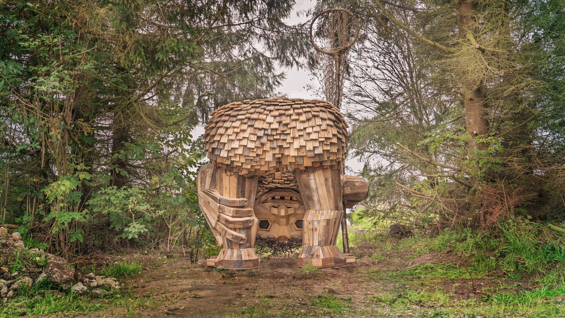 Large wooden sculpture resembling a bear, built from stacked rectangular planks, standing on four blocky legs in a forest clearing. The open mouth and brown wood contrast with surrounding green trees.