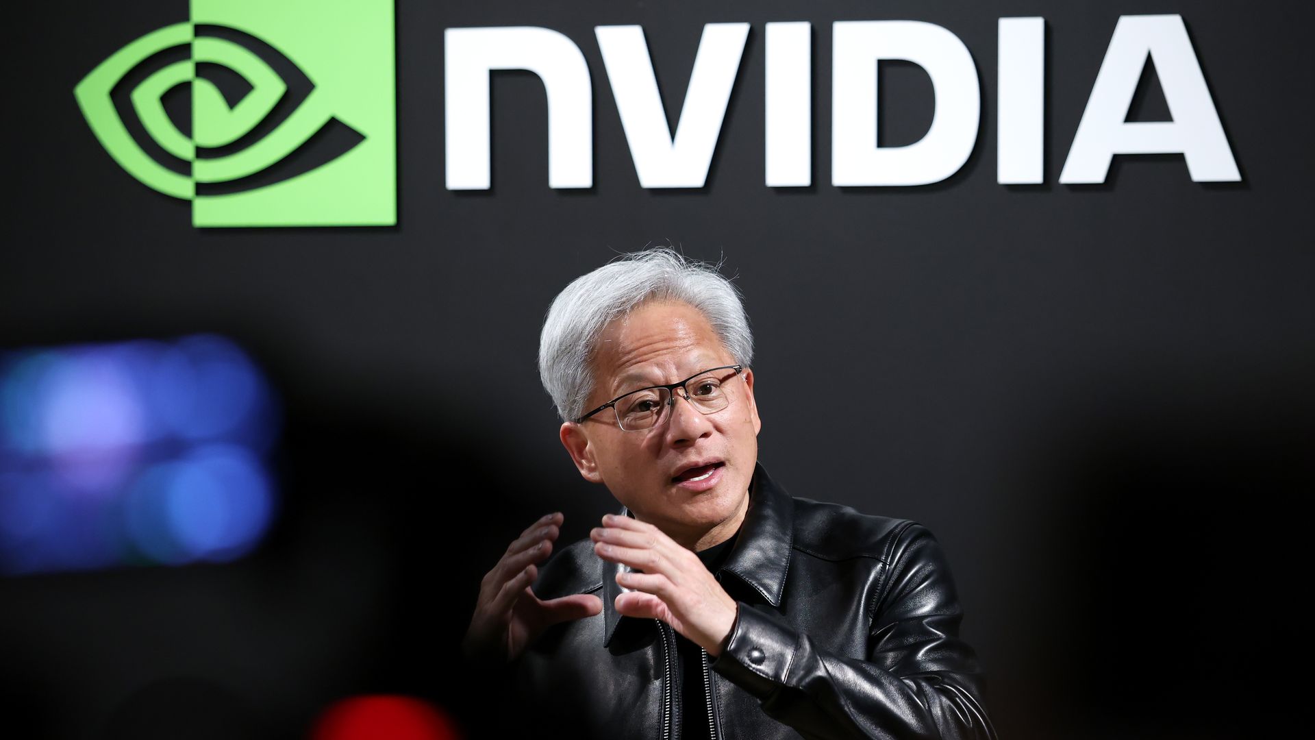 A person with gray hair and glasses wearing a black leather jacket speaks, gesturing in front of a large NVIDIA sign with a green square eye logo on a dark wall.