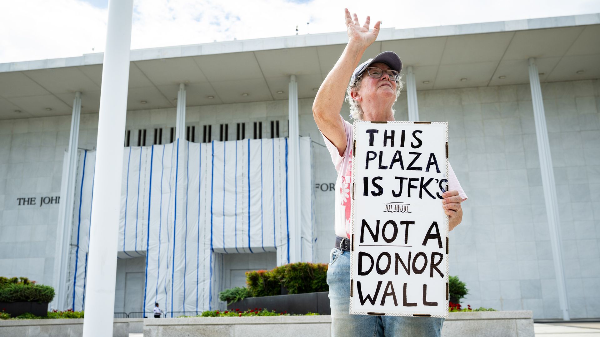 A demonstrator — a woman wearing a hat, jeans and glasses — stands outside of the Kennedy Center, the front of which is covered by tarp, while she waves and she holds a sign that reads "THIS PLAZA IS JFK'S NOT A DONOR WALL."