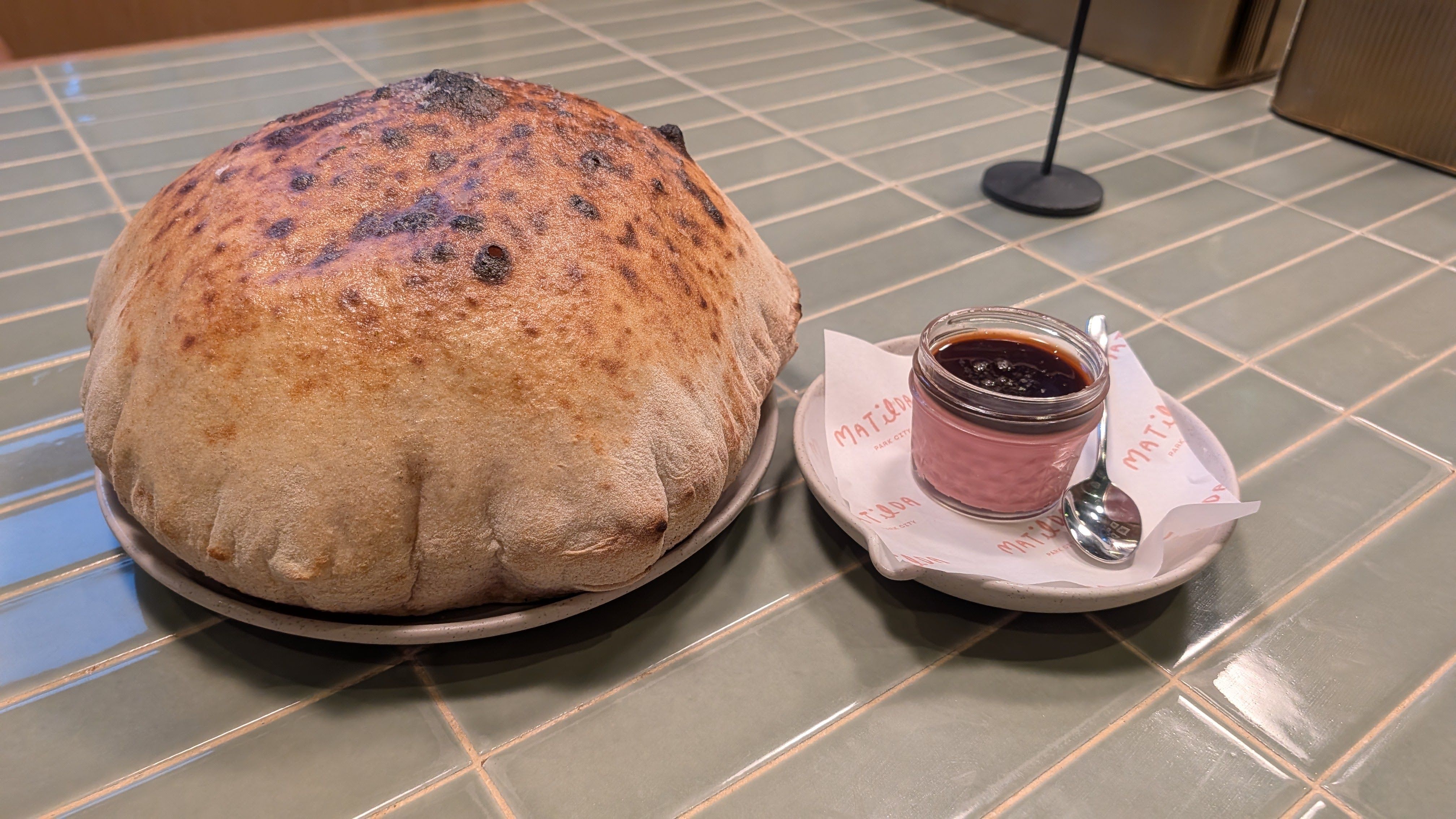 Large, round, crusty loaf on a plate beside a pink-tinted jar of dark purple jam, with a napkin and spoon on a pale green tiled counter.