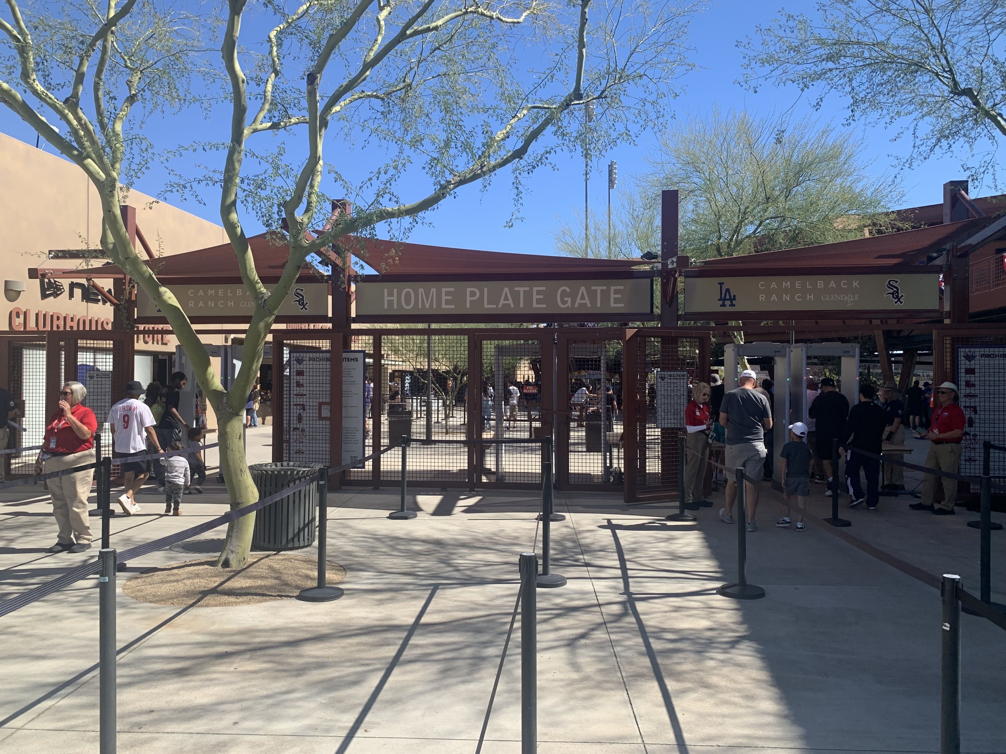 Entrance gate labeled Home Plate Gate at Camelback Ranch Glendale baseball stadium with people entering, metal detectors, trees, and clear blue sky.