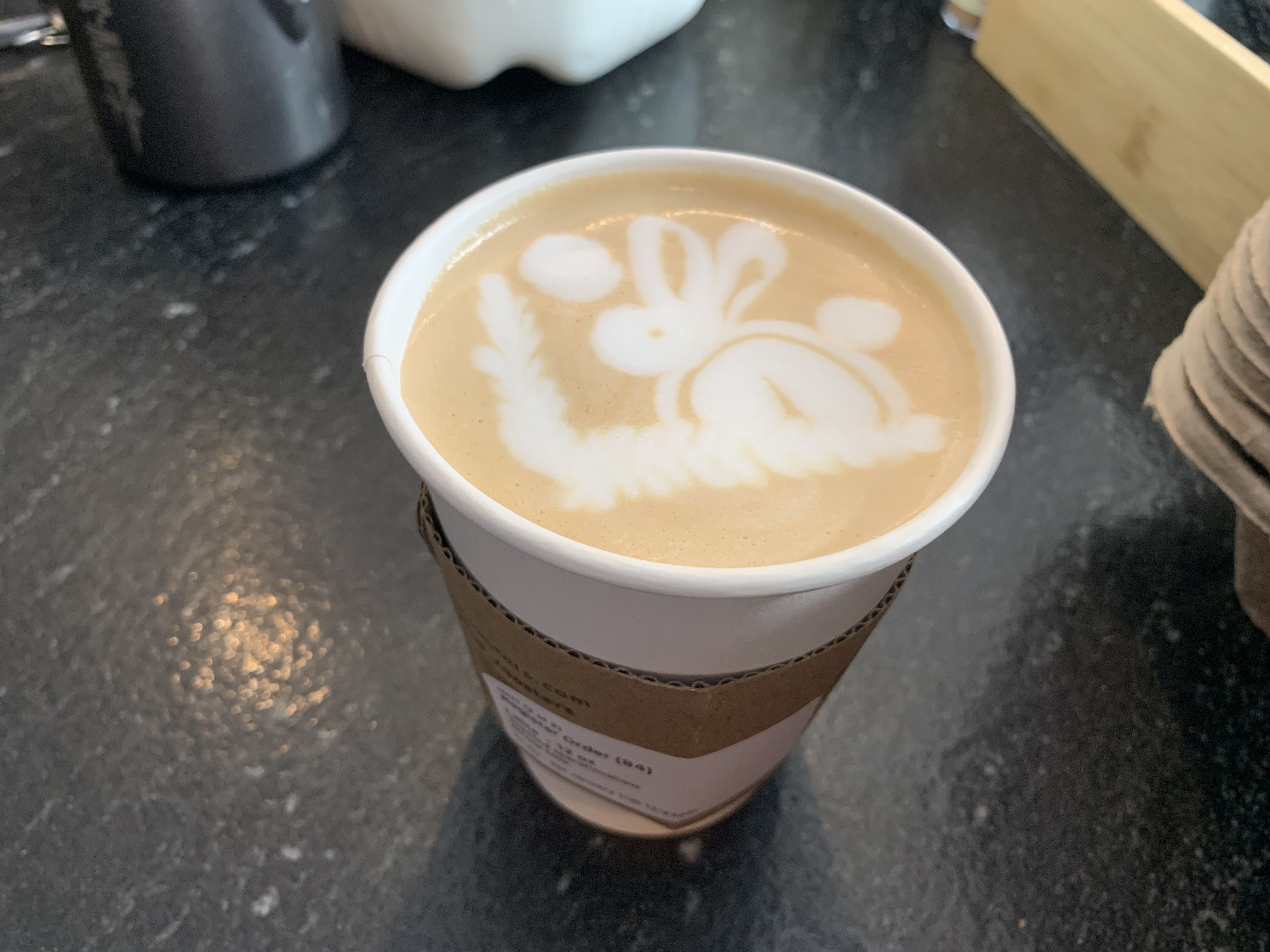 Paper cup of latte with foam art depicting a rabbit with ears and a clock, placed on a dark countertop with various objects blurred in the background.