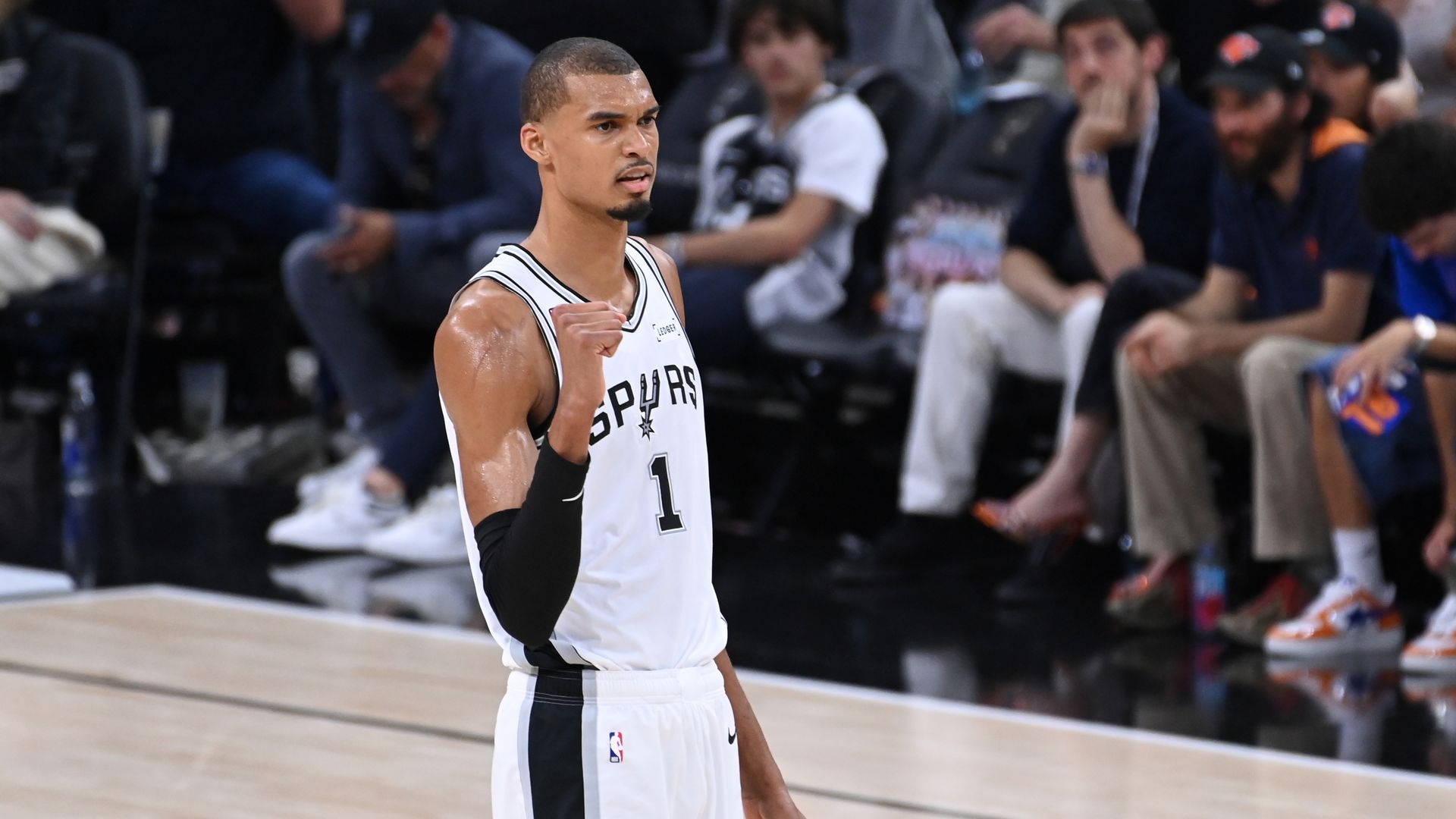 A Spurs basketball player in a white jersey with the number 1 raises a clenched fist on the court, with a blurred crowd in the background.