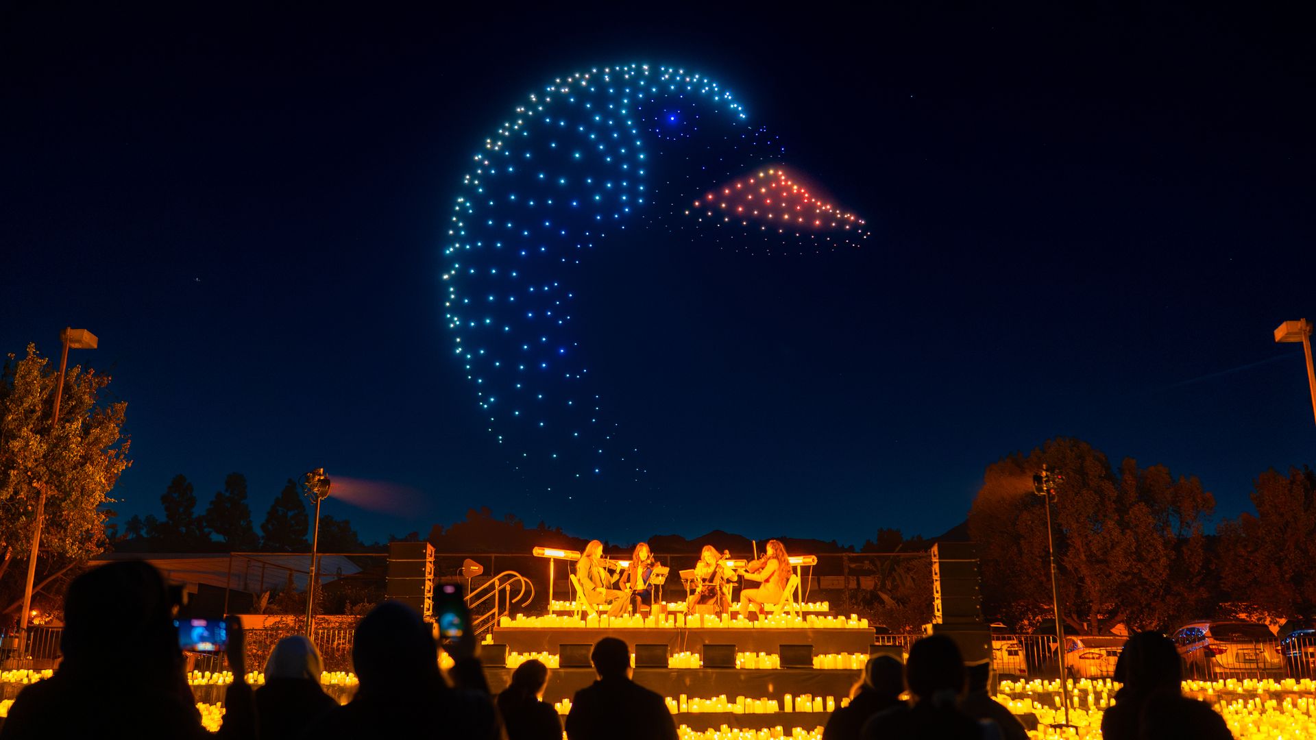 A string quartet performing on a candle-lit stage outdoors with an illuminated large bird during a dark night. 