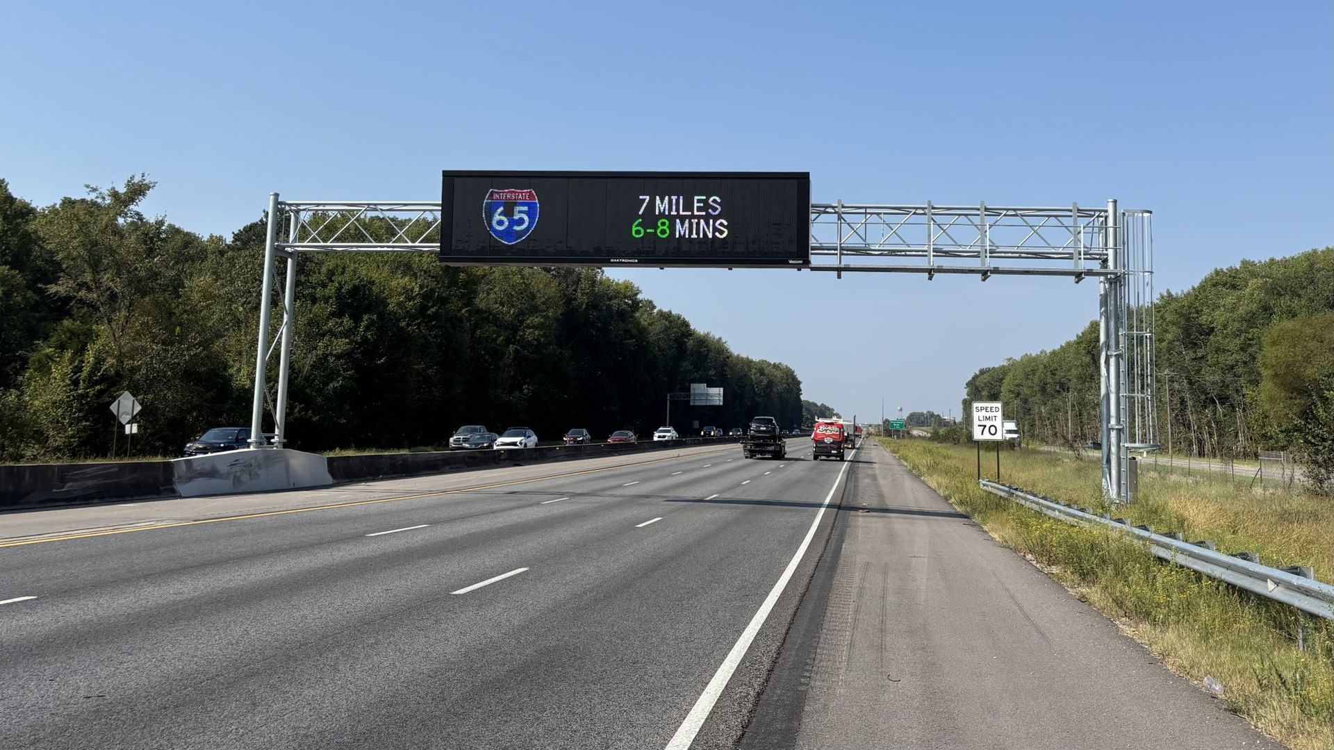 Highway scene on a clear day with a digital sign above indicating Interstate 65 is 7 miles away, travel time 6-8 minutes. Speed limit sign shows 70 mph on the right side.