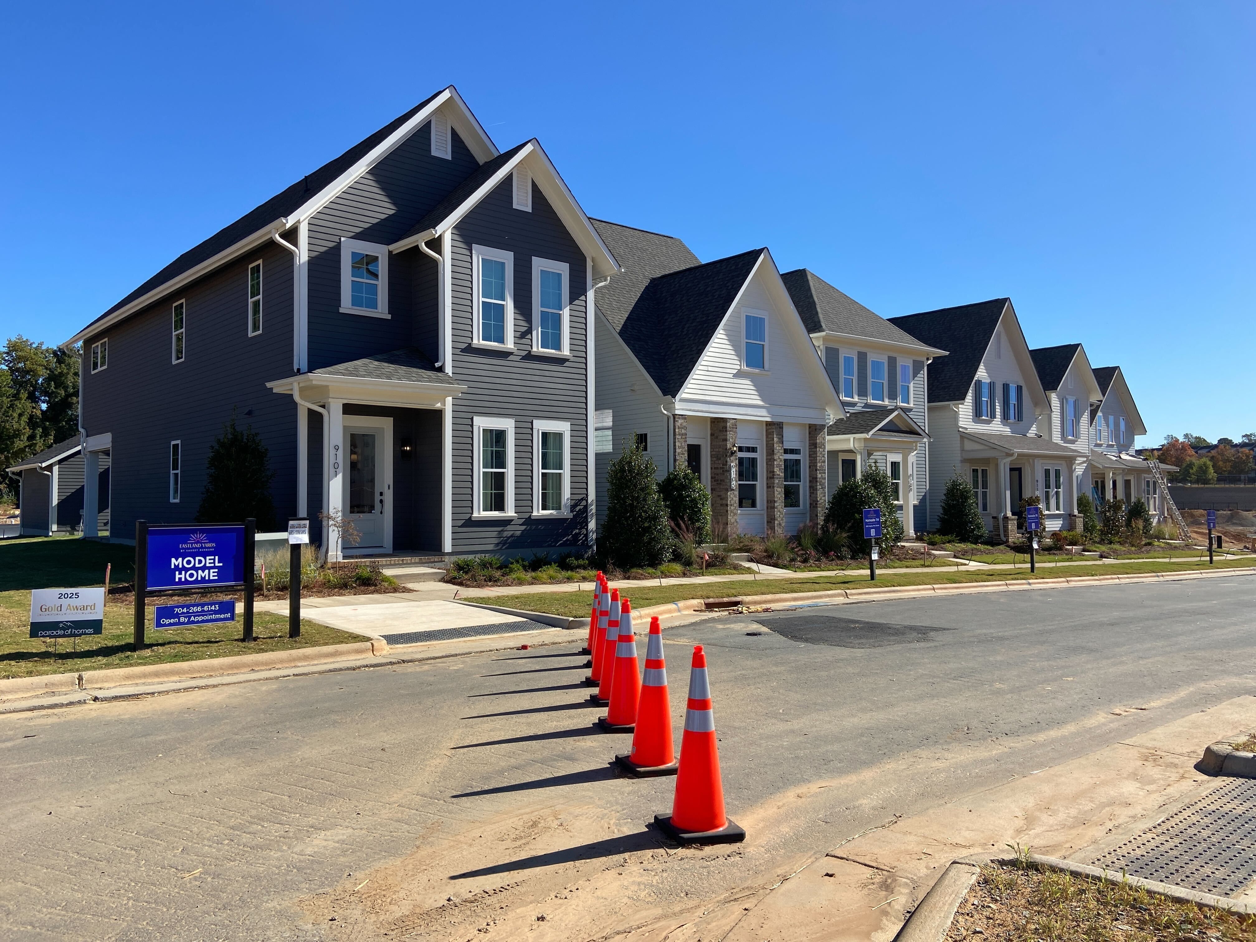 Row of suburban model homes with gray and white siding under clear blue sky, orange traffic cones line the street blocking access, and signs indicating the model home and awards.