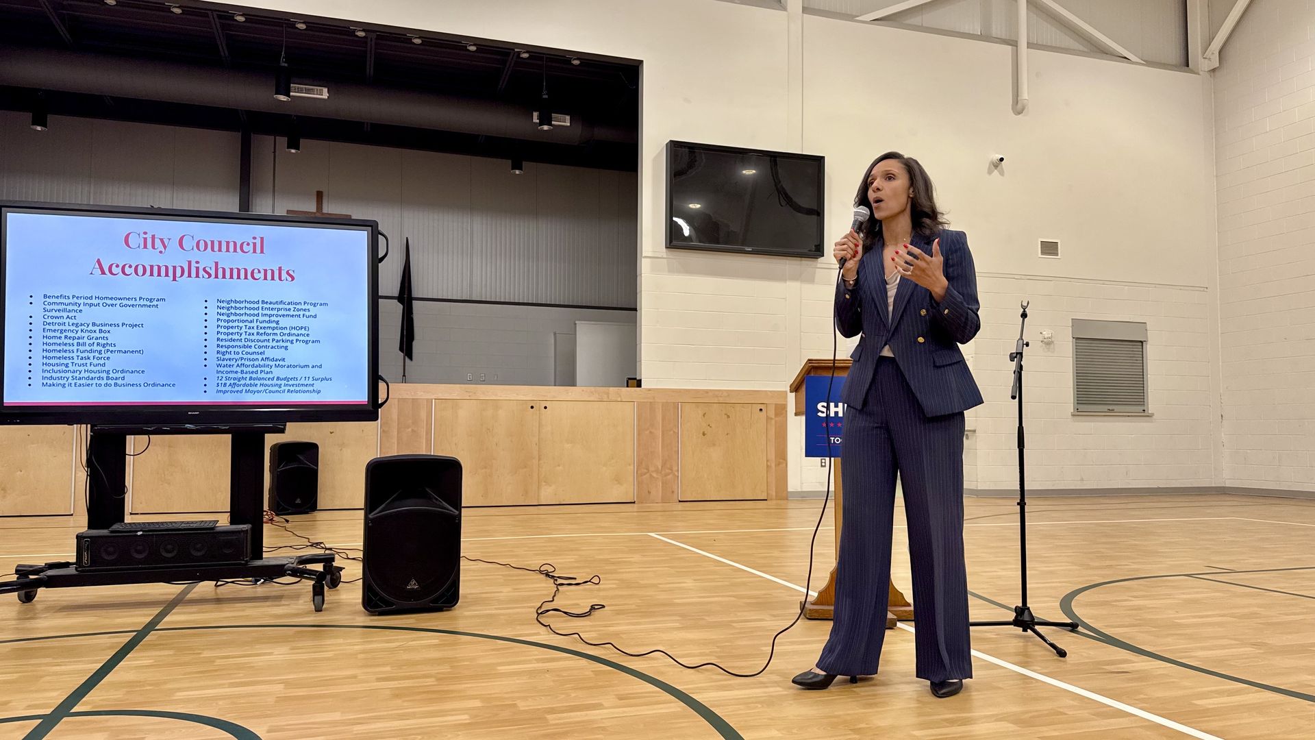 Sheffield, wearing a suit, speaking into a microphone in a gymnasium with a screen nearby displaying City Council accomplishments.