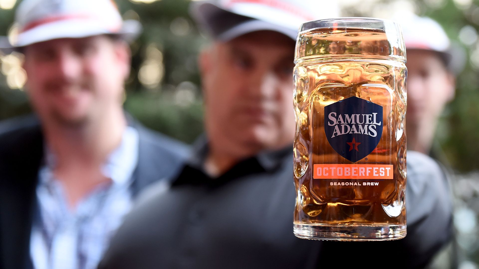A close up on a stein with the Samuel Adams Oktoberfest beer logo with three men in the background.