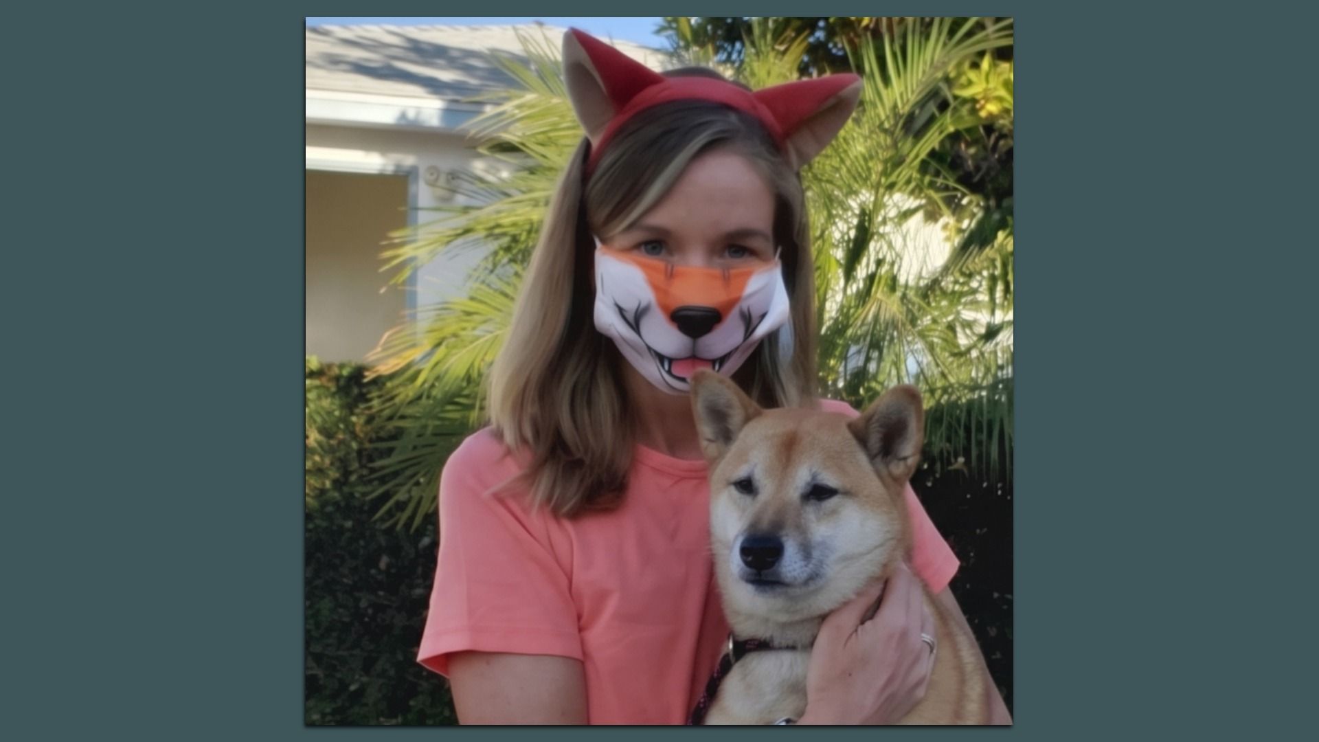 Person with blonde hair wearing a red fox-ear headband and a fox-face mask in a pink shirt, holding a tan Shiba Inu outdoors, with palm plants and a house in the background.