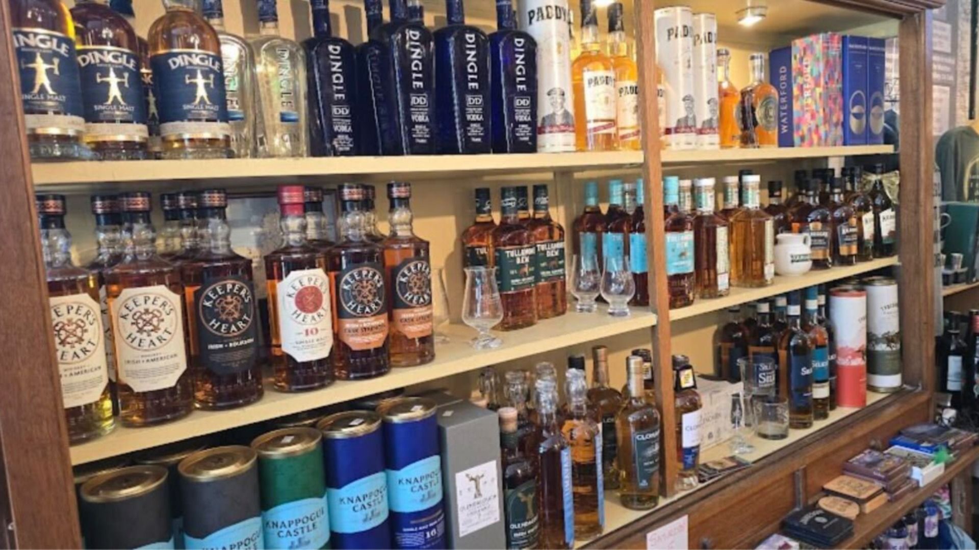 Wooden liquor store shelf crowded with whiskey bottles across three levels; visible labels include Dingle and Keeper's Heart, plus boxed bottles, tumblers, and assorted packaging.