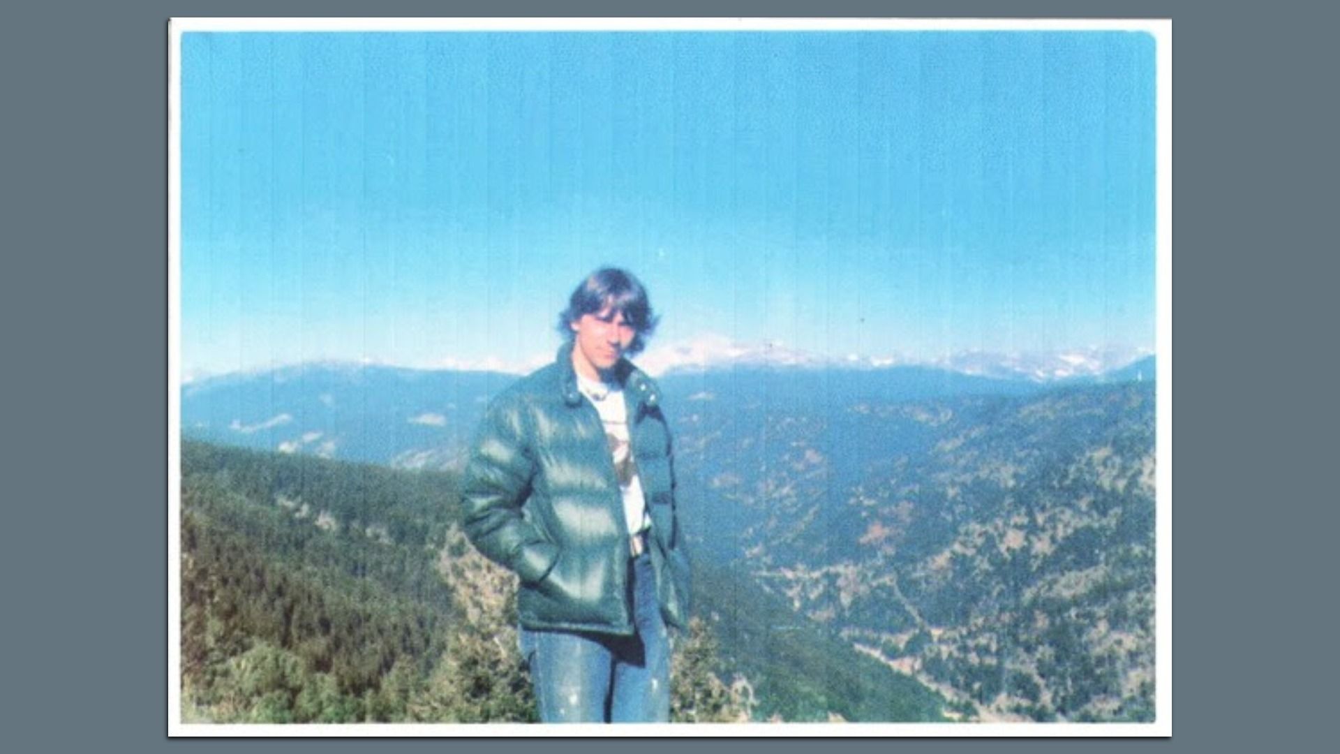 John Curtis Patterson stands in front of a mountain range wearing jeans and a jacket in an older photo.