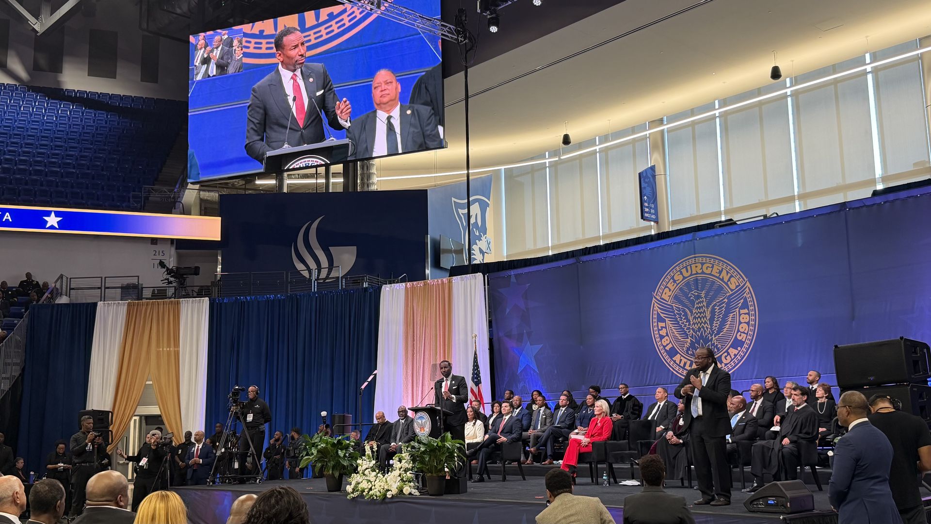 Man in black suit and red tie speaking at a podium on stage with seated audience and sign language interpreter, large screen above and Atlanta city seal on blue backdrop.