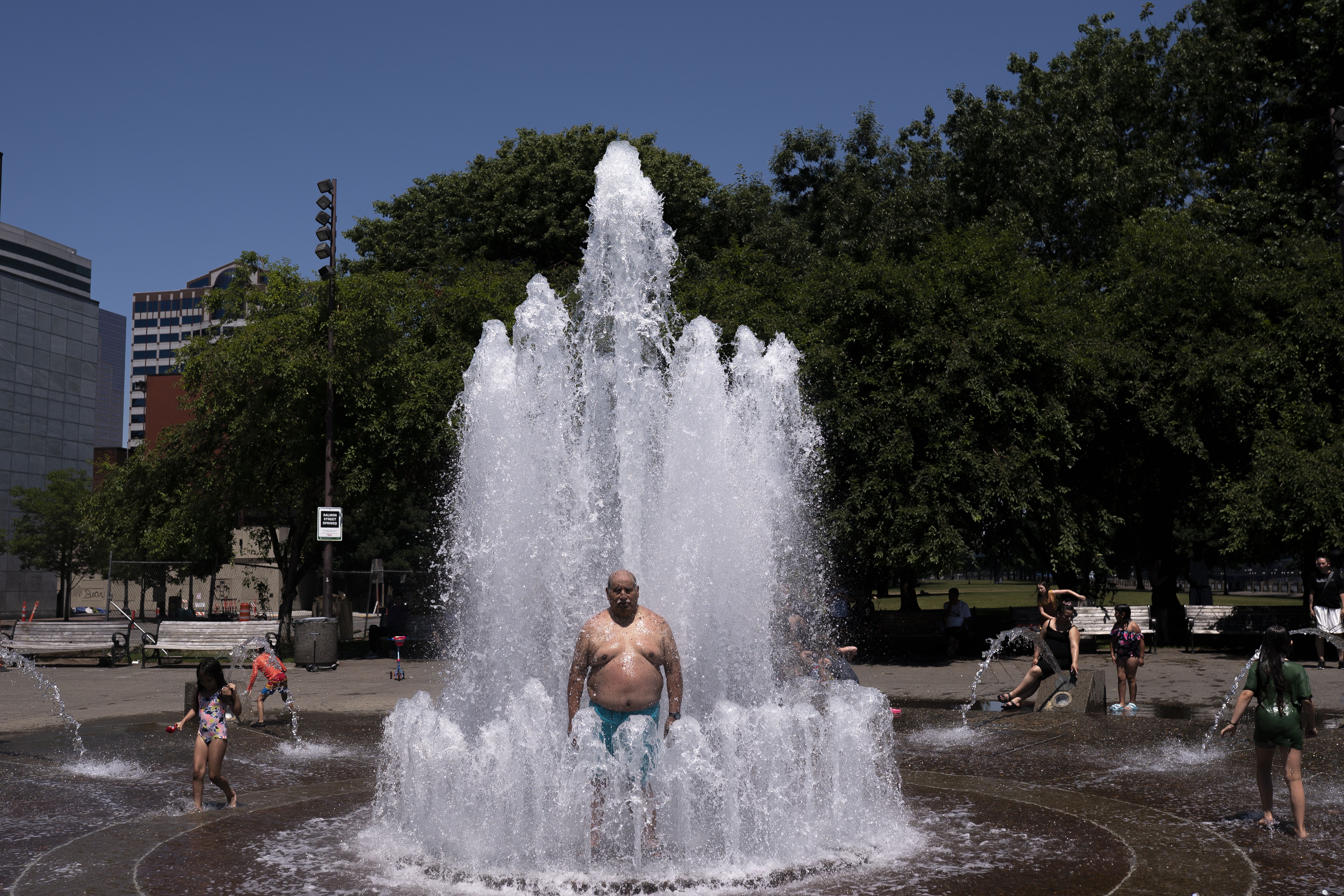 A large city fountain shoots water upward in a plaza. A shirtless man in teal shorts stands in the spray as children play nearby, with green trees and tall buildings behind under a clear blue sky.