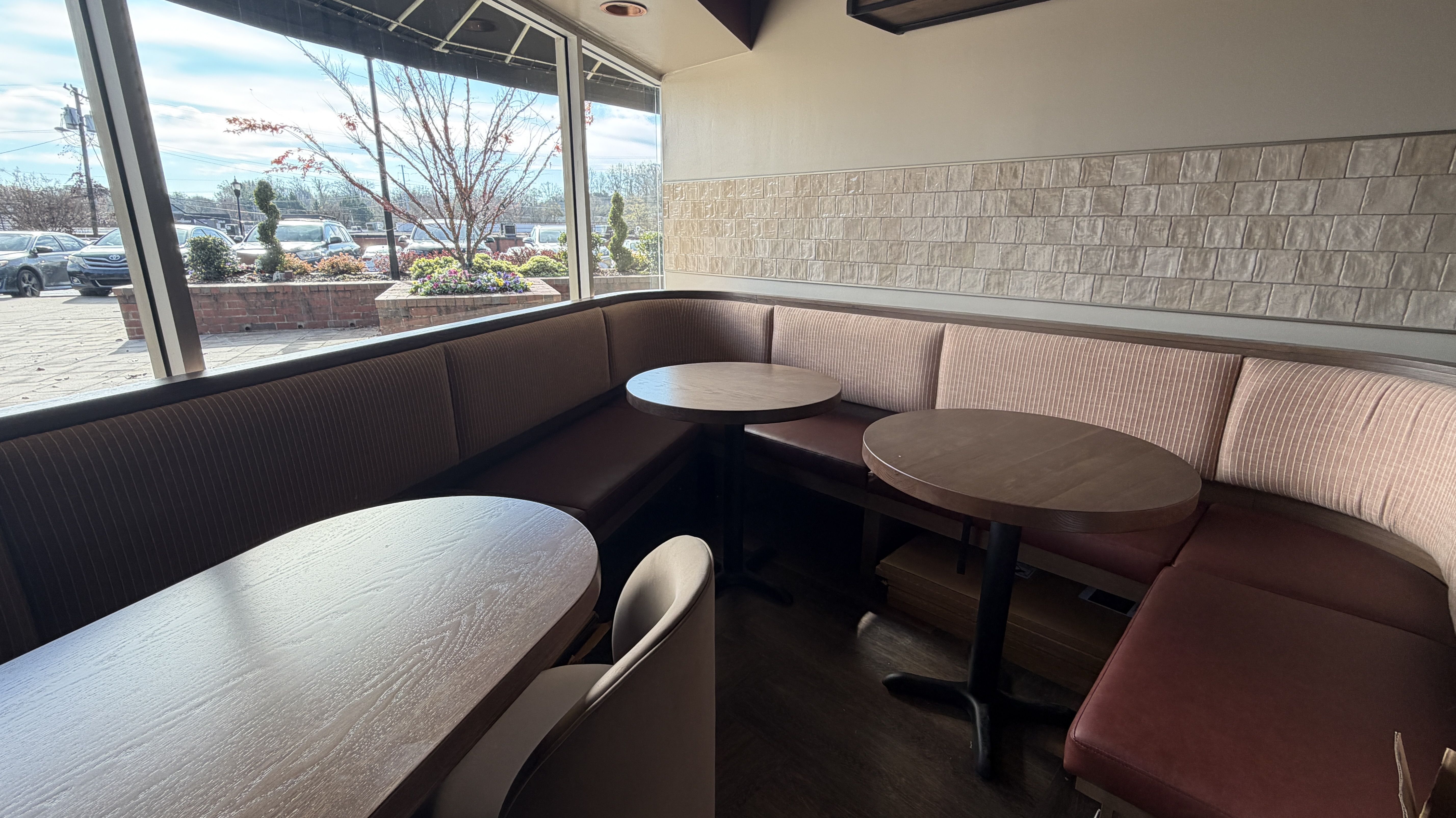Cozy restaurant corner with brown curved bench seating, two small round wooden tables, one oval wooden table, a single beige chair, large windows showing cars and trees outside.