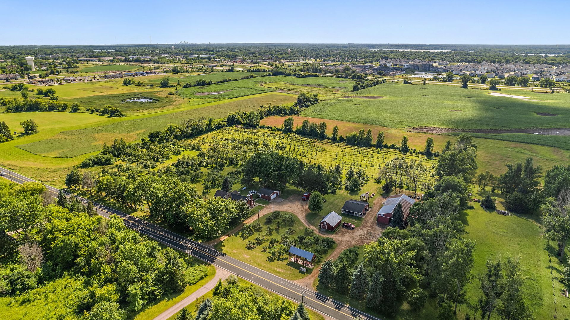 Aerial view of a green farm with several red buildings, fields, and trees under a clear blue sky, with roads and a suburban neighborhood visible in the distance.
