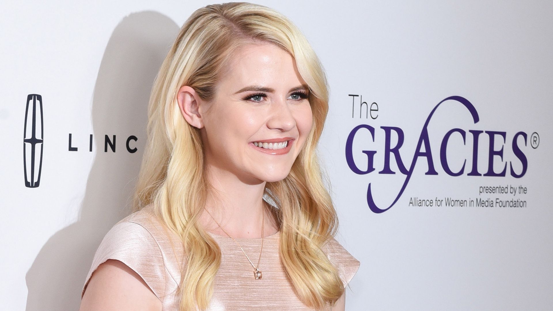 Blonde woman smiling on a red carpet, wearing a pale pink satin dress. She stands in front of a step-and-repeat backdrop with a large purple GRACIES logo and sponsor text.