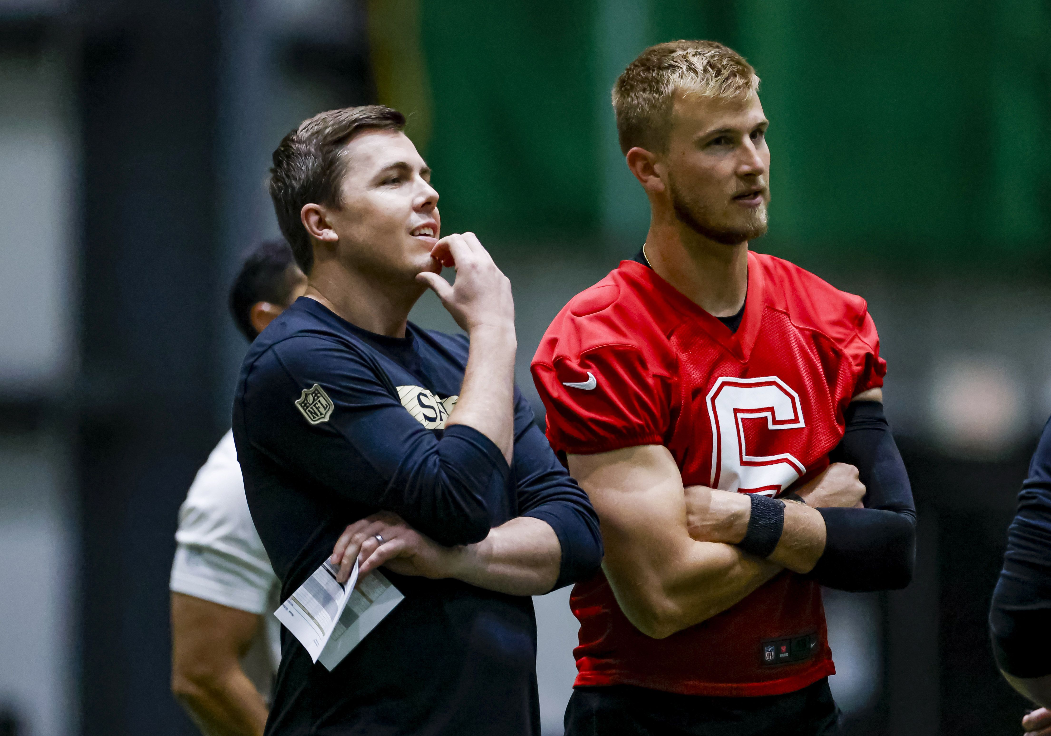 Two men stand side by side indoors; one wears a black NFL shirt holding a paper, the other a red sports jersey with number 6, both looking focused and engaged in thought.