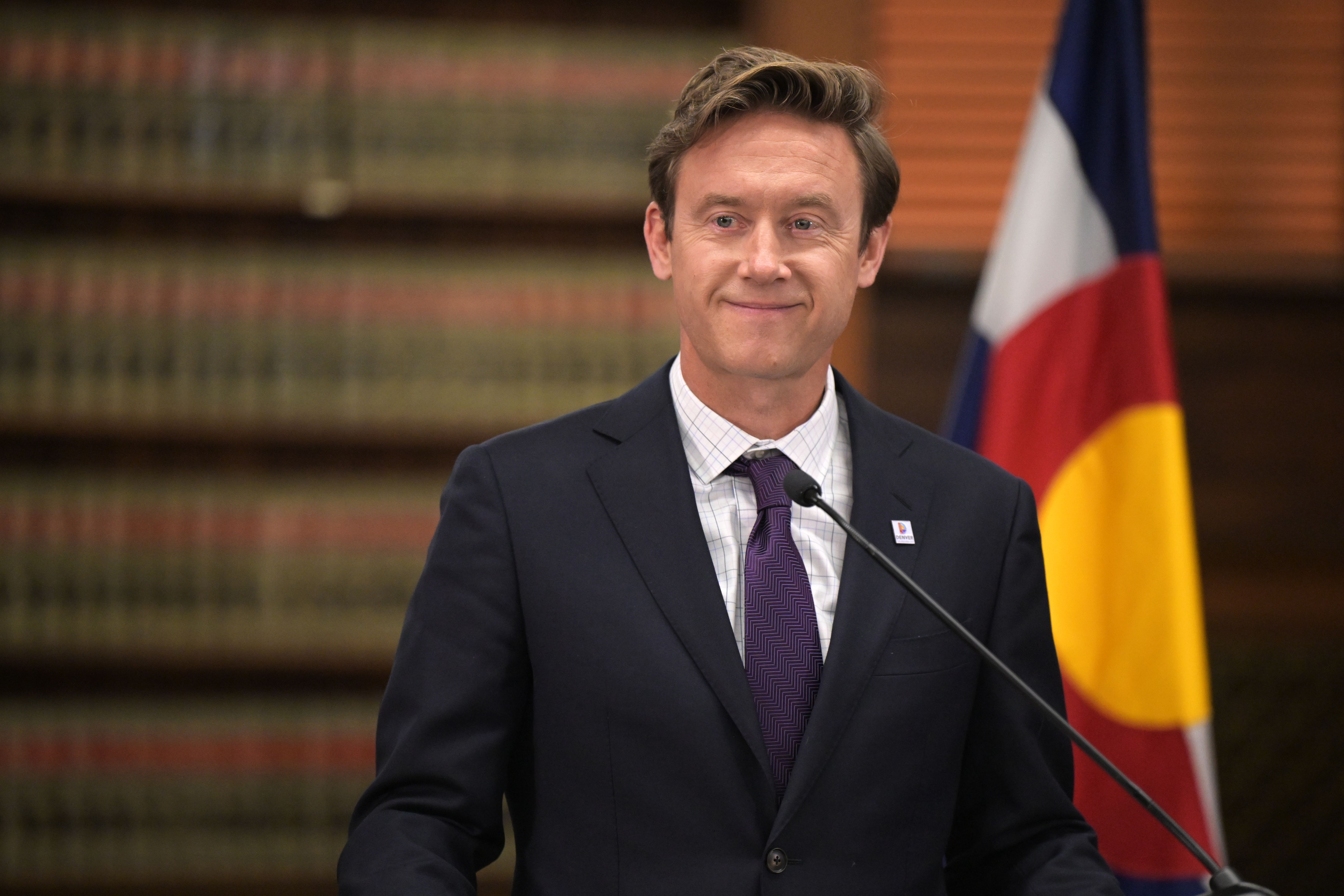 A white man in a suit in front of a flag