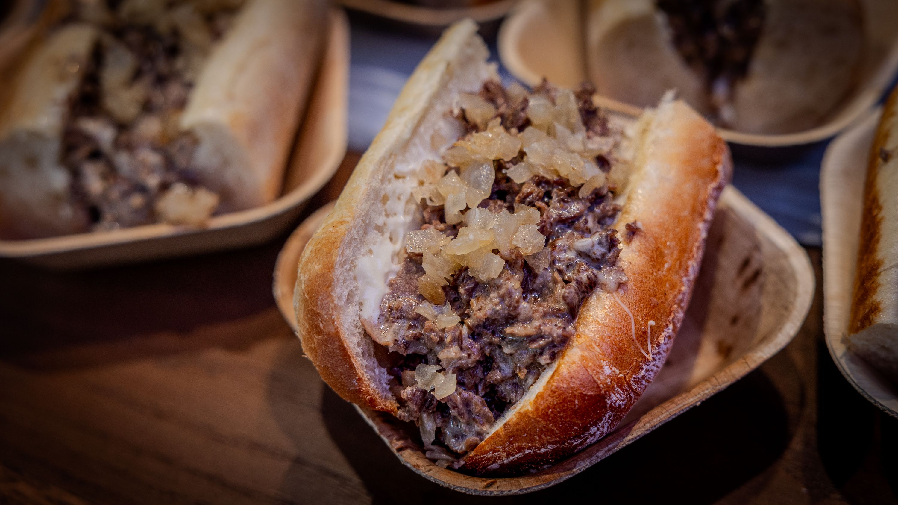 Close-up of a sandwich with chopped beef and diced onions in a toasted bun, served in a small brown paper tray on a wooden surface.
