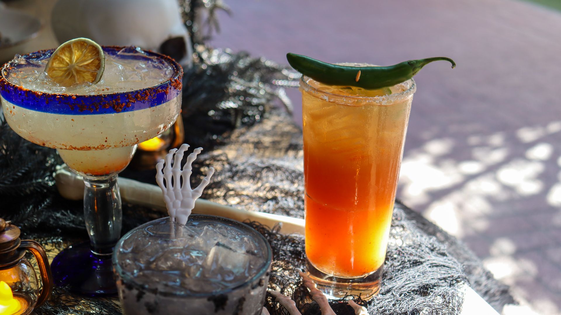 Two cocktails on a festive table: a frosted margarita with a lime wheel and a blue rim, and a tall orange gradient drink topped with a green chili pepper garnish.