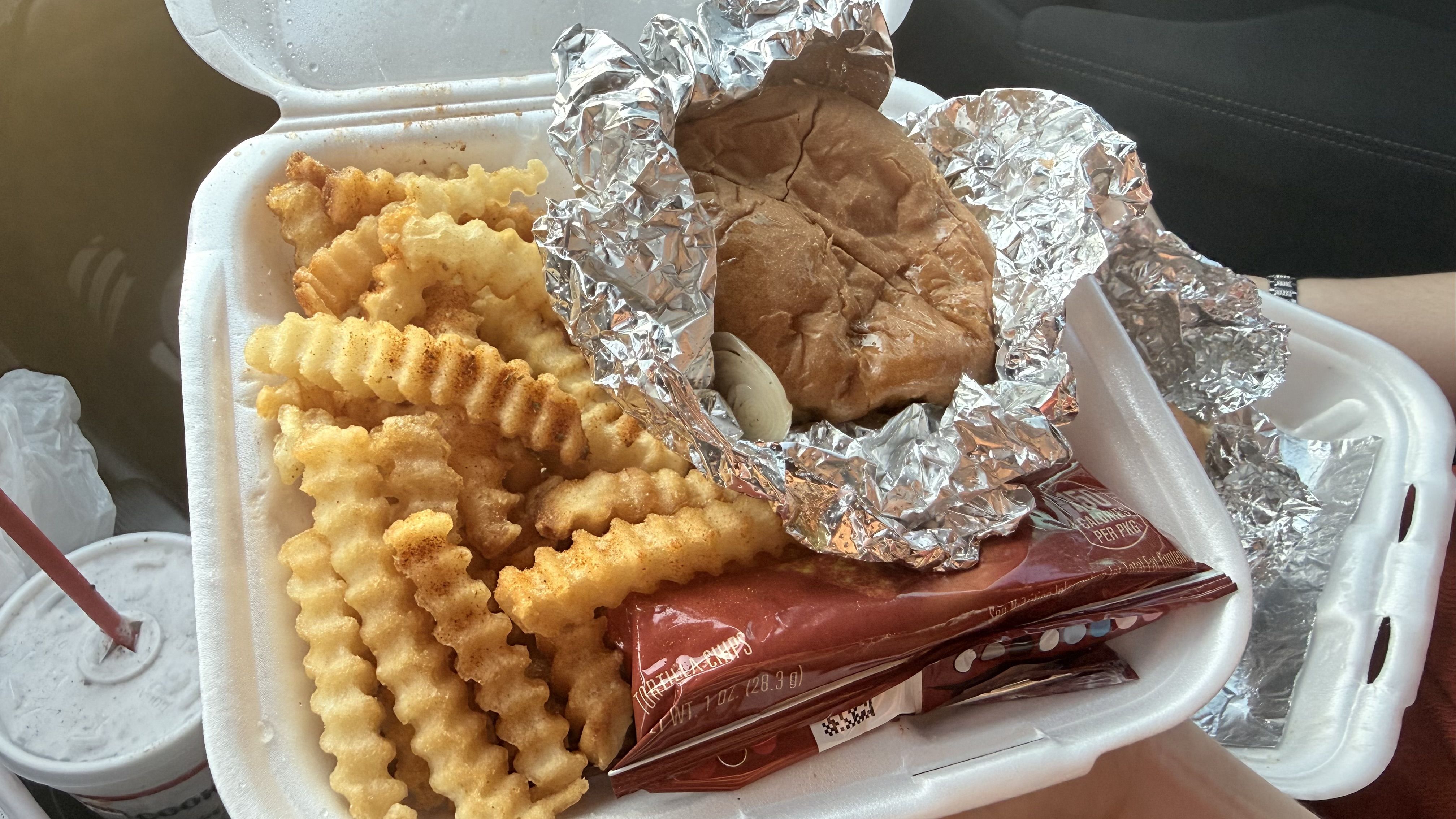 Open foam takeout box with golden crinkle-cut fries and a foil-wrapped sandwich in a brown bun; a red straw and crumpled foil wrappers nearby.