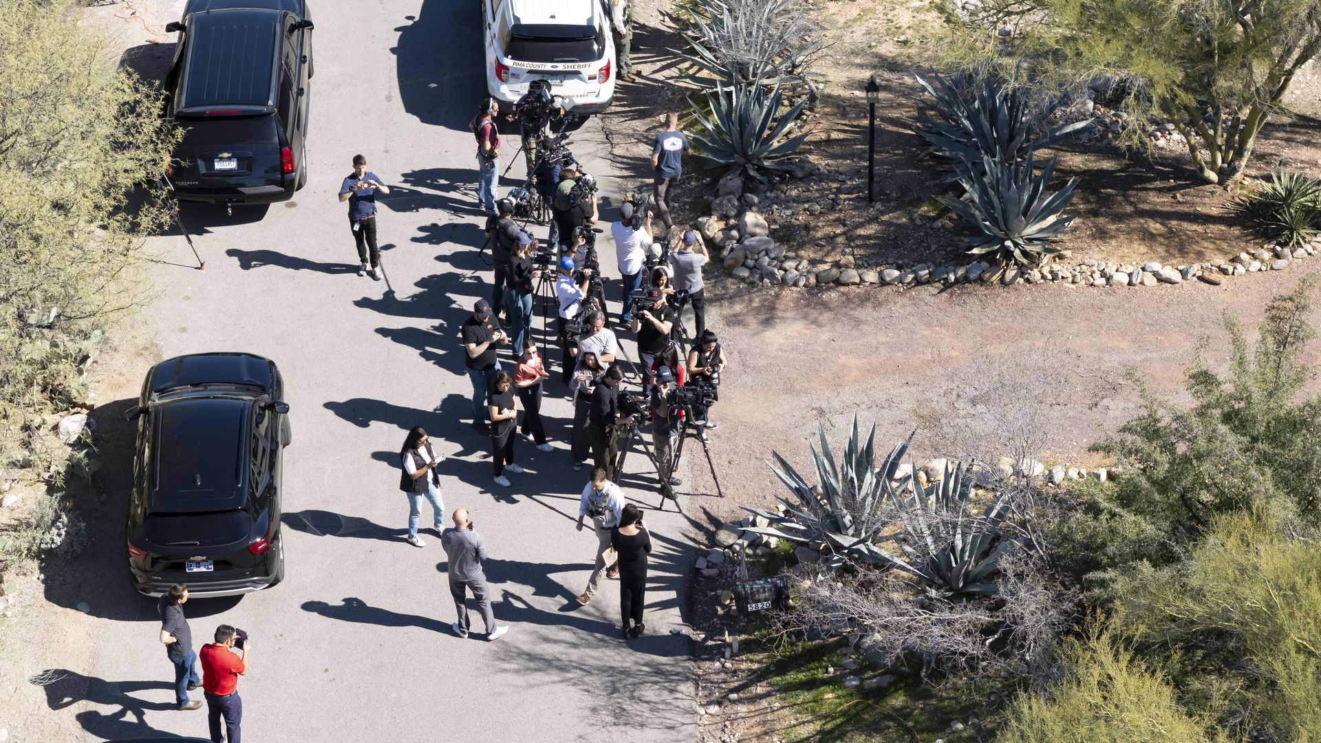 A group of photographers and reporters with cameras and tripods on a sunny street near desert plants, alongside several parked black SUVs and a Pima County Sheriff's vehicle.