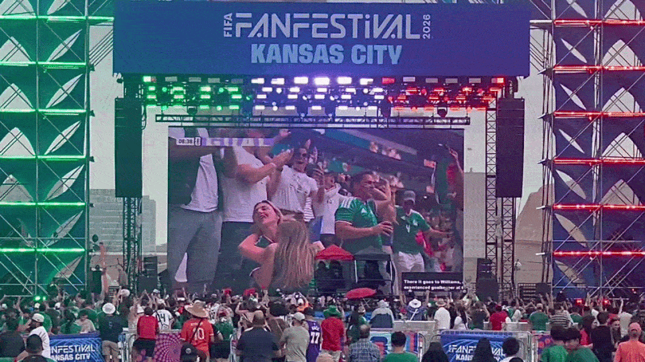Outdoor festival stage with a giant LED screen and multicolored lighting; a crowded audience, many in green jerseys, watches the show. Banner above reads "FAN FESTIVAL KANSAS CITY 2026".