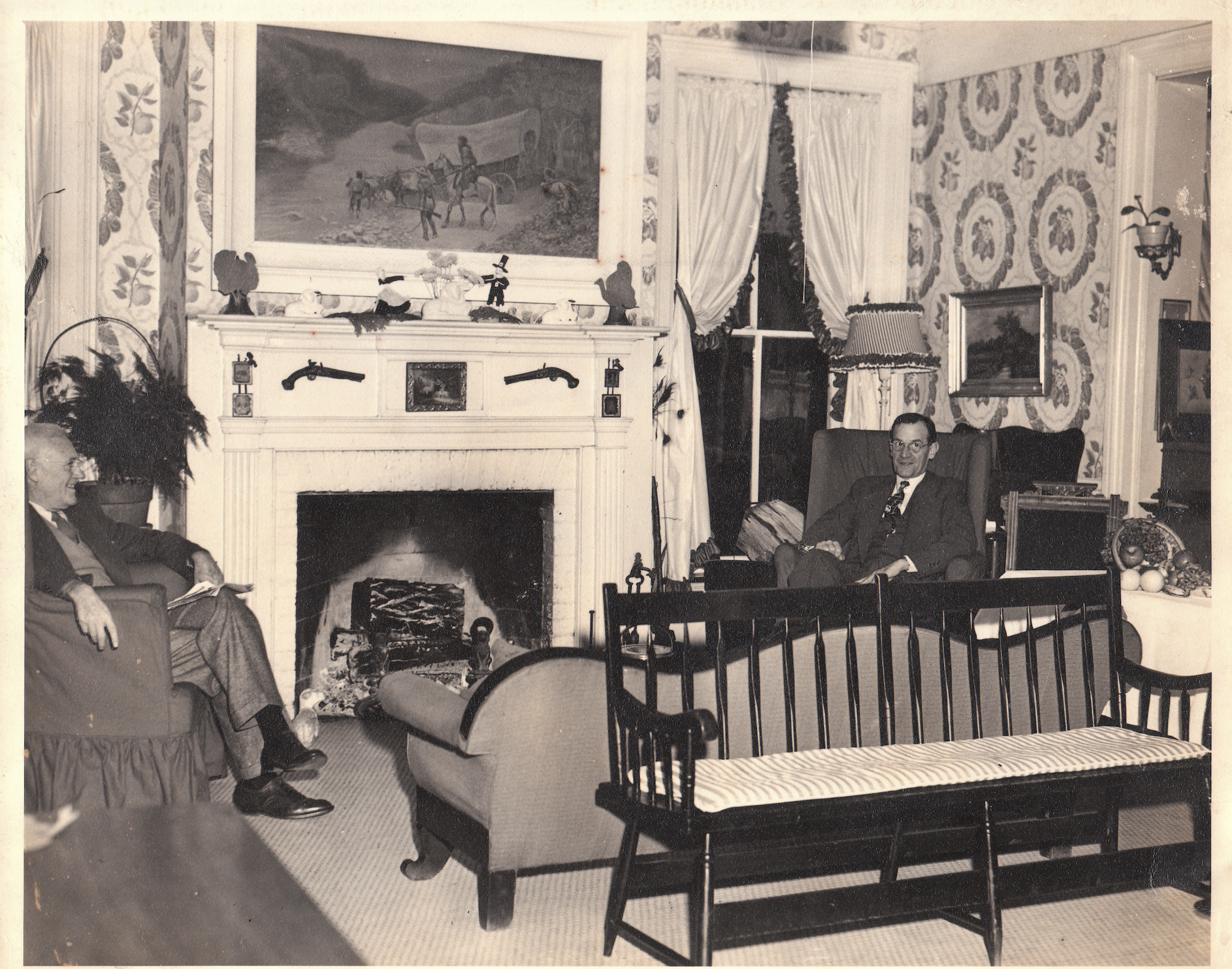 Black-and-white photo of a vintage living room with wallpaper, white mantel, a wagon-train painting, heavy curtains, two men seated left and right, and a baby crib in the foreground.