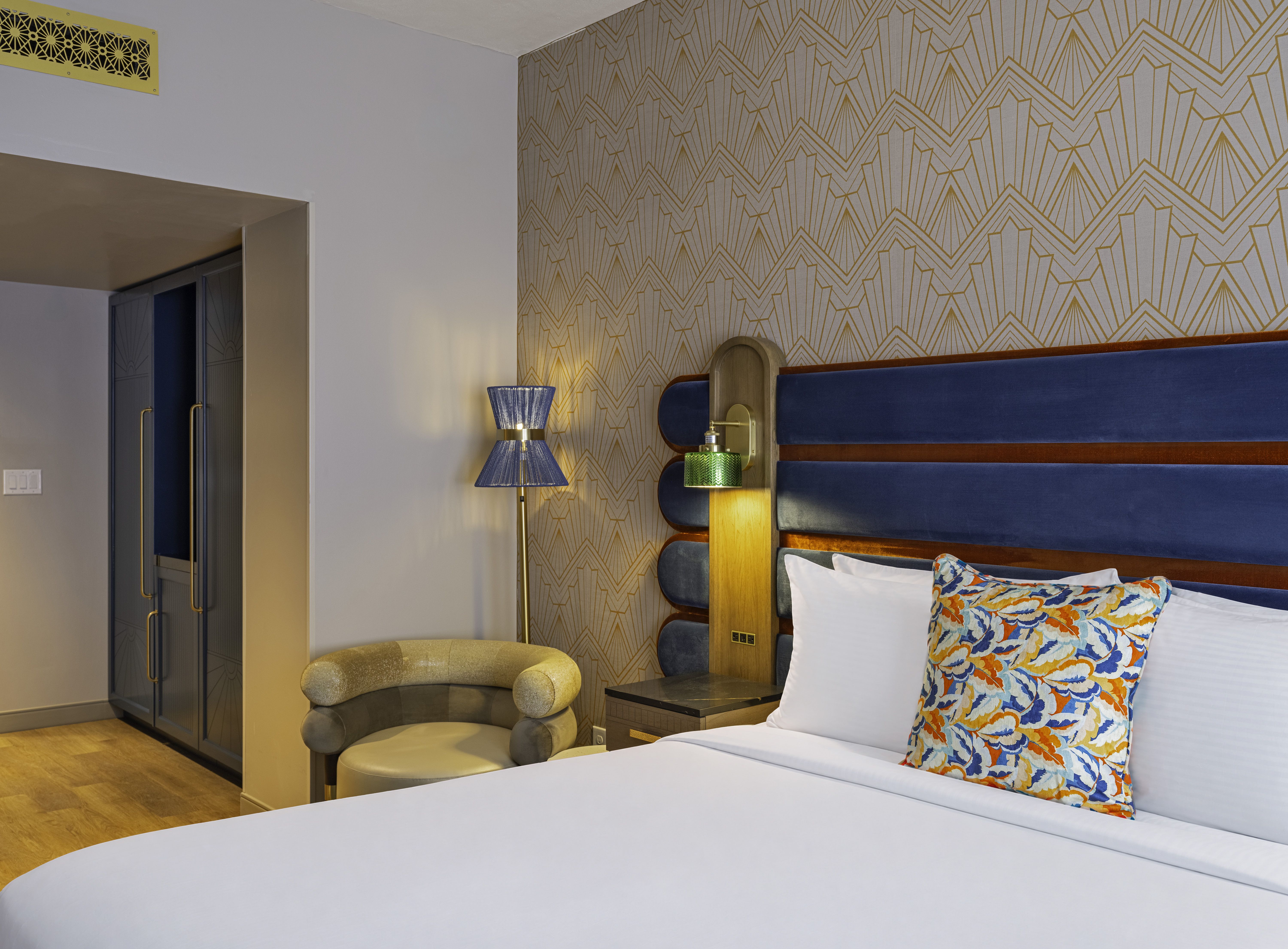 Hotel room with a tall blue headboard, gold geometric wallpaper, and nightstands. A blue lampshade and a green glass lamp light the bed with floral pillows; doorway on the left.