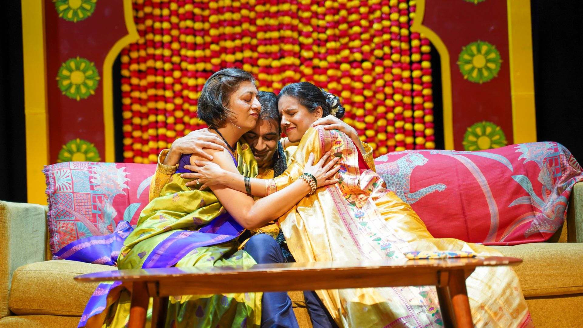 Two women in bright sarees hug and smile on a man stage, with a vivid red-yellow floral backdrop and a patterned pink sofa in front.