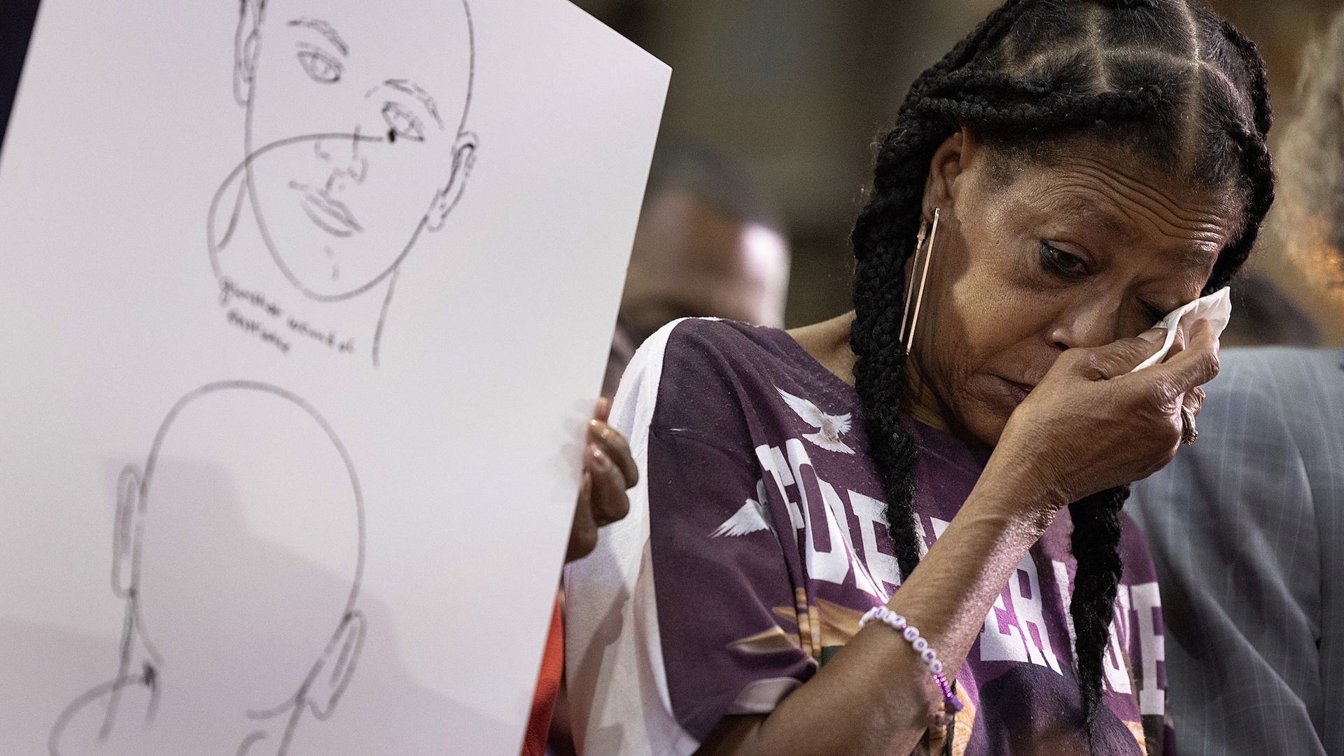 A woman with braids and gold earrings wipes tears away with a tissue standing next to a sketch of a face with the direction of a bullet.