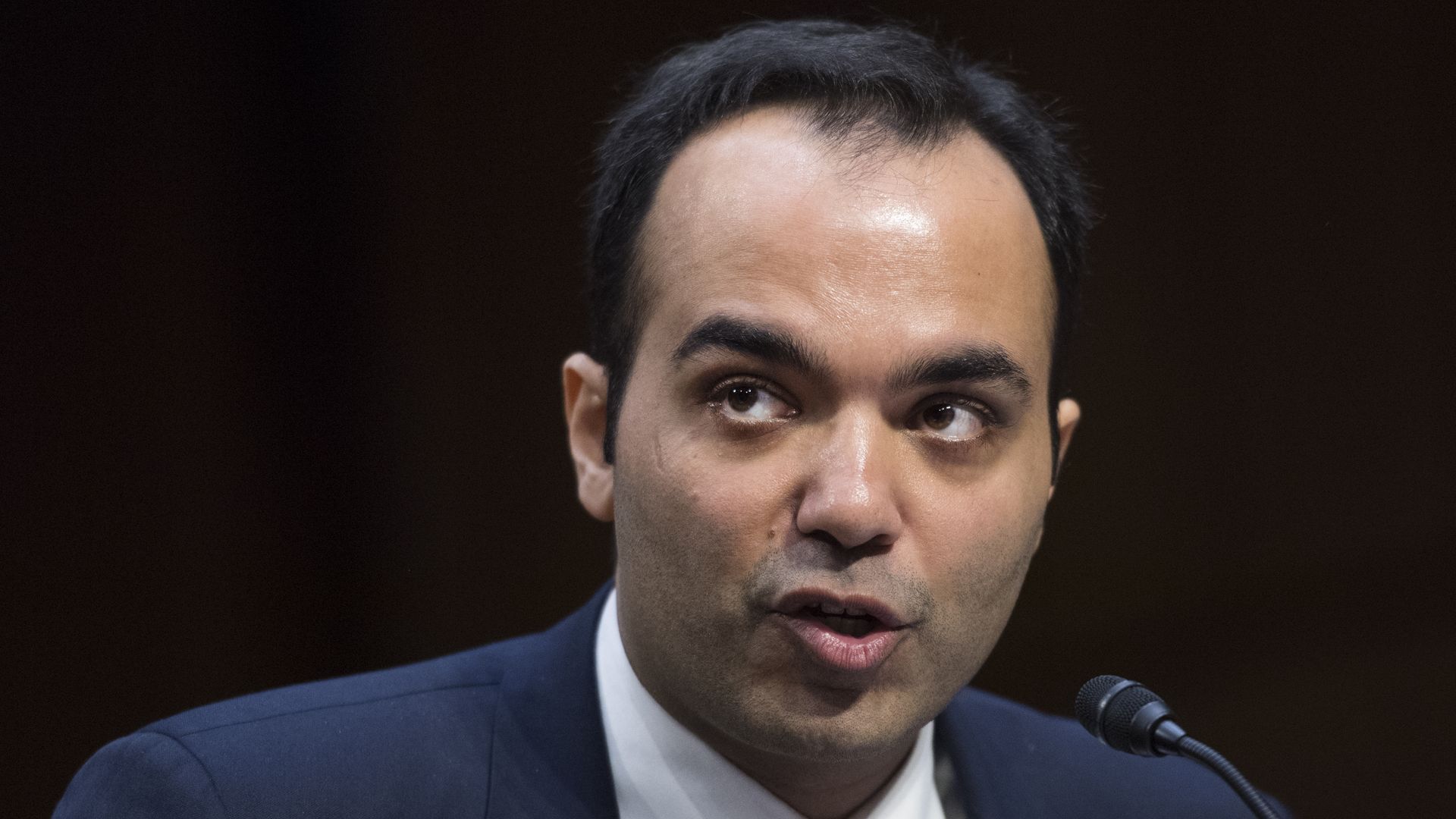 FTC Commissioner Rohit Chopra speaks at a microphone, wearing a suit and tie