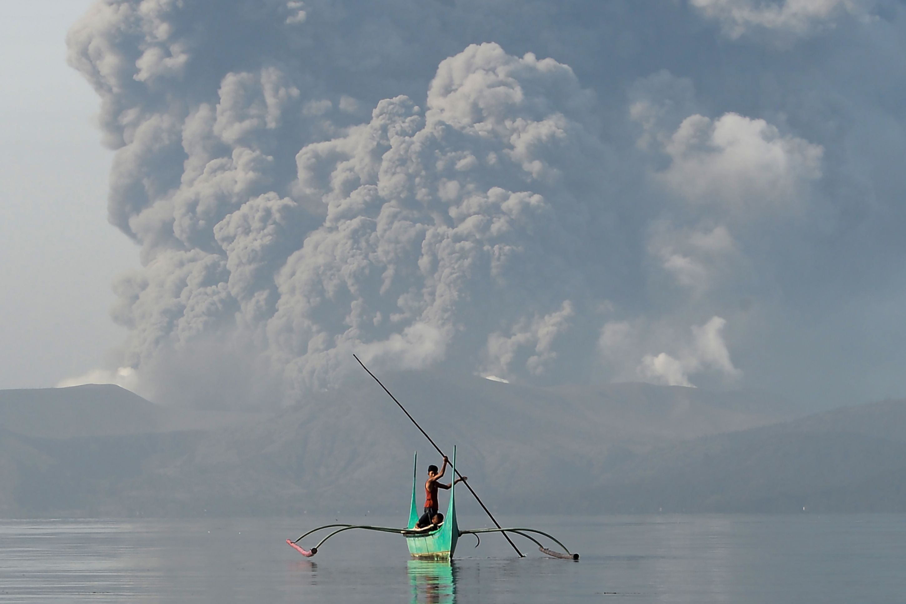 A youth living at the foot of Taal volcano rides an outrigger canoe while the volcano spews ash as seen from Tanauan town in Batangas province, south of Manila