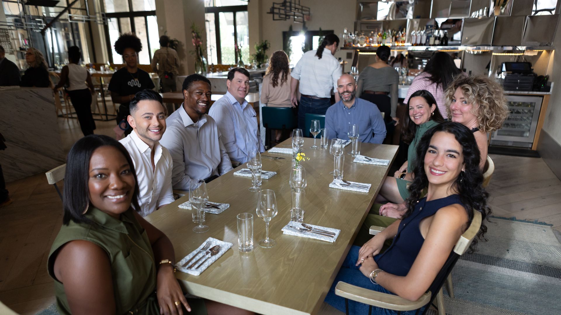 Diverse group of people sits around a long wooden dining table in a bright restaurant, smiling at the camera; the table has wine glasses, cutlery, napkins, and a small yellow flower centerpiece.